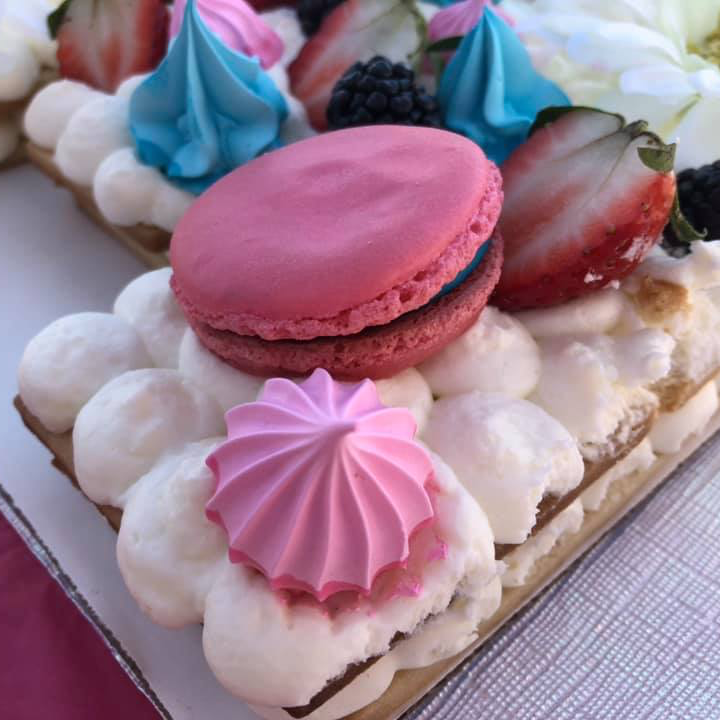 A rectangular pastry topped with white cream, pink and blue meringue swirls, a pink macaron, strawberries, and blackberries.