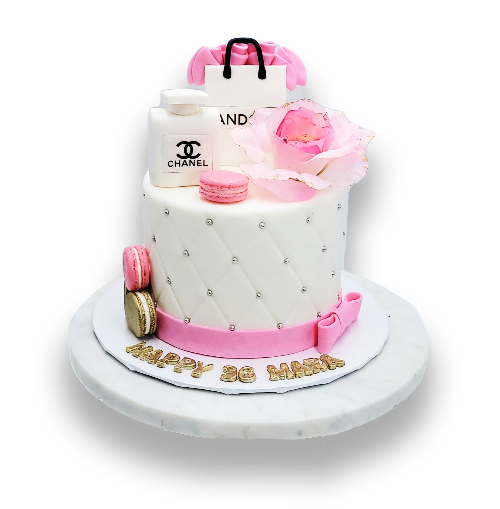 A white quilted cake topped with a Chanel-themed bag, perfume bottle, pink flower, and macaroons on a gold-lettered base.