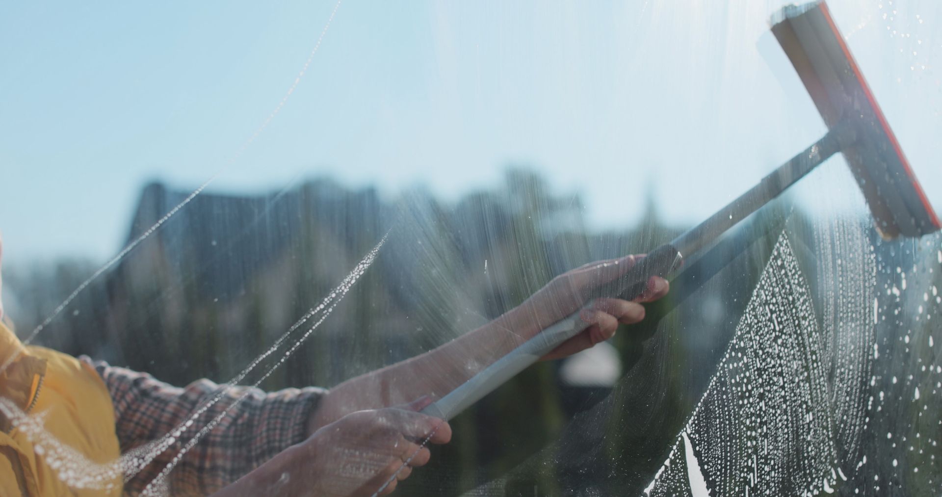 Person washing a window with a squeegee in bright sunlight, water spraying across the glass.