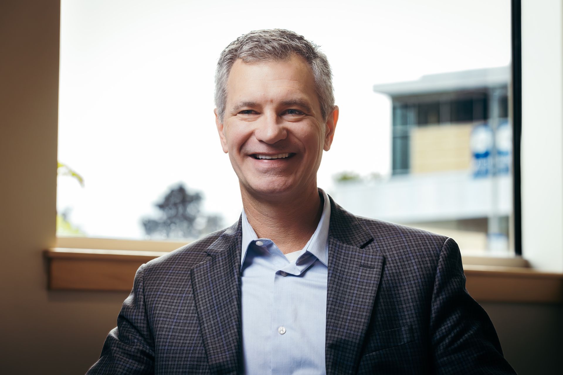 A man in a suit and blue shirt is smiling while sitting in front of a window.
