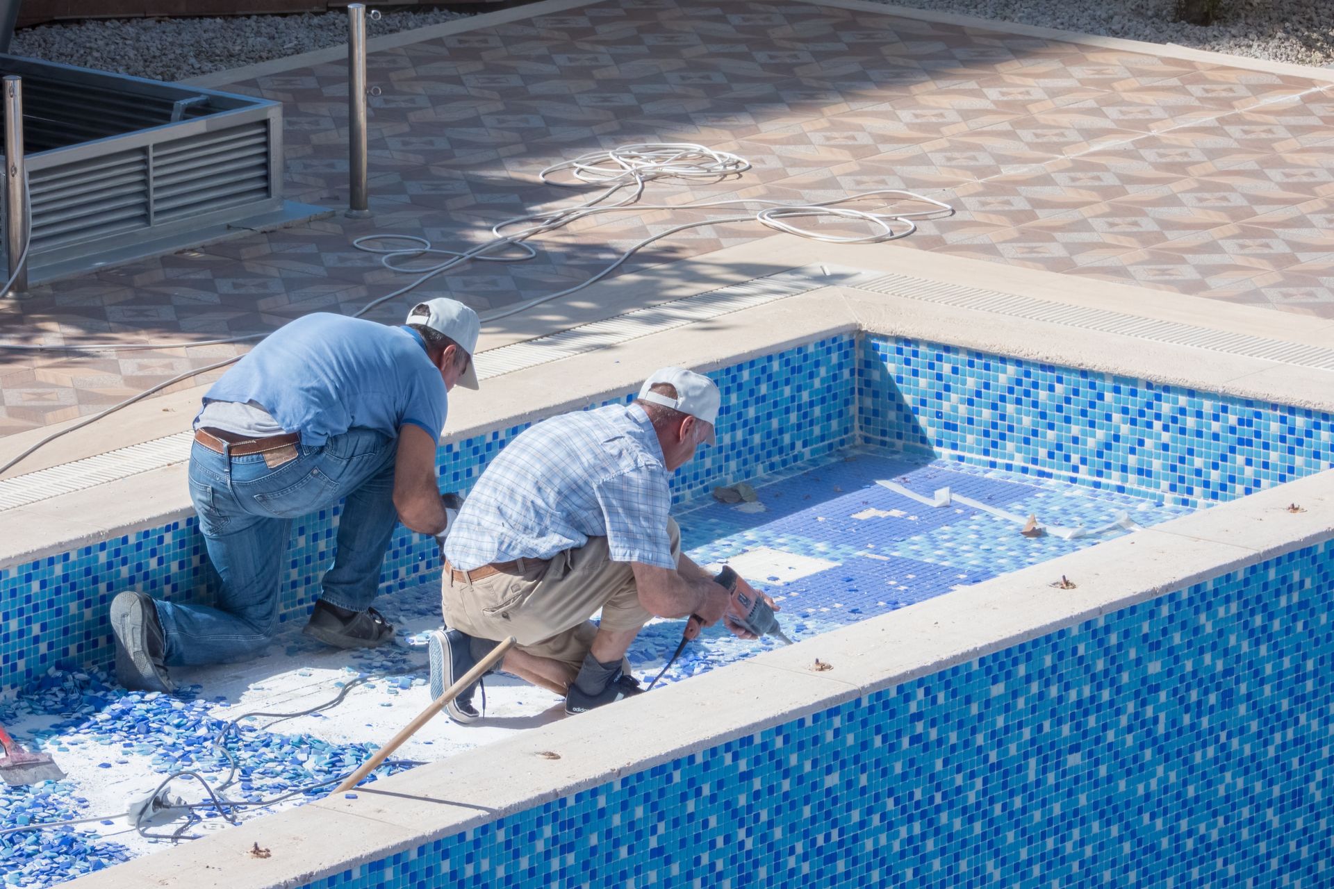 Two men removing tiles from a blue tiled swimming pool. They wear hats and work outside.