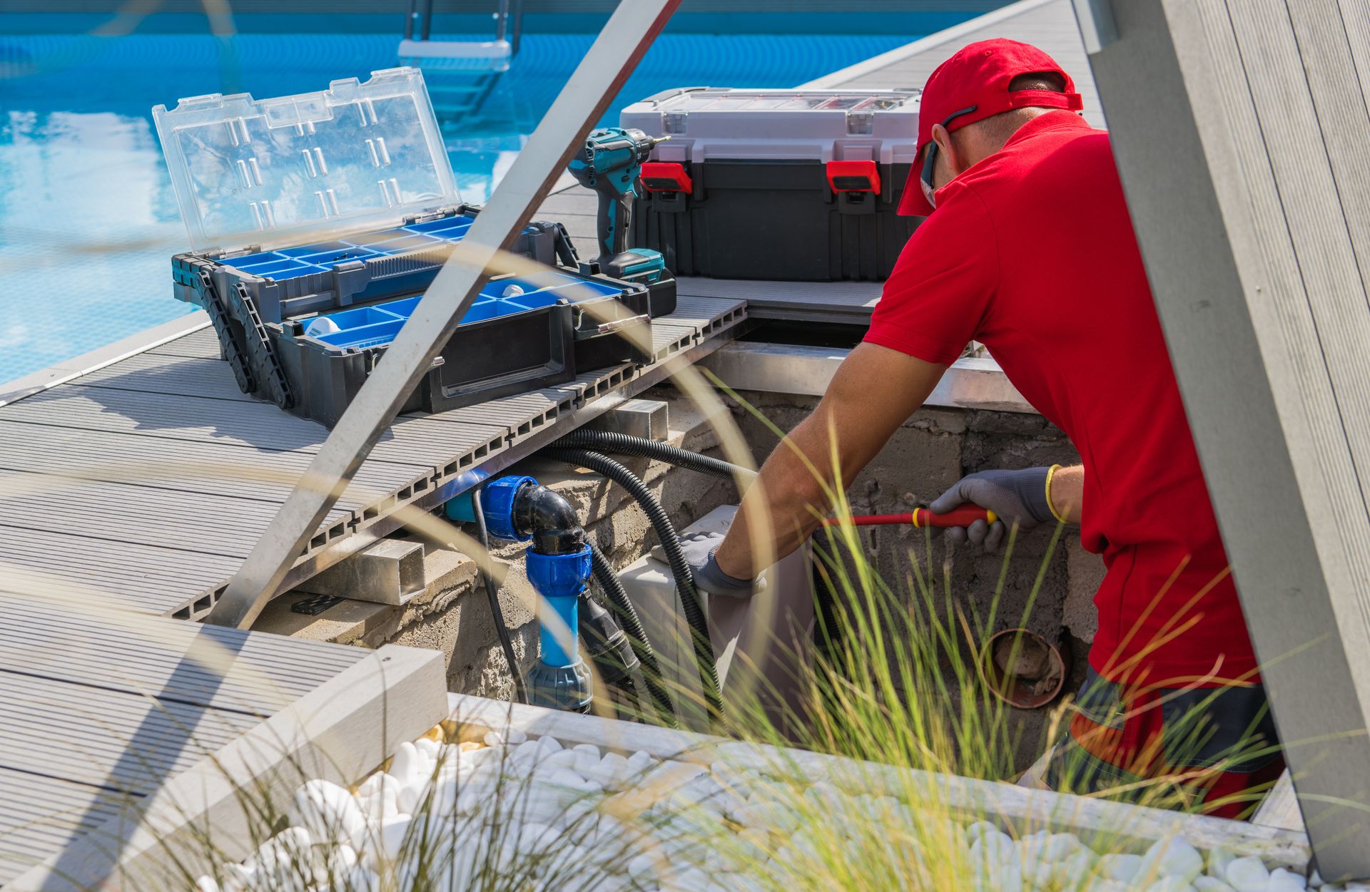 Pool technician in red shirt repairing pool equipment next to a swimming pool.