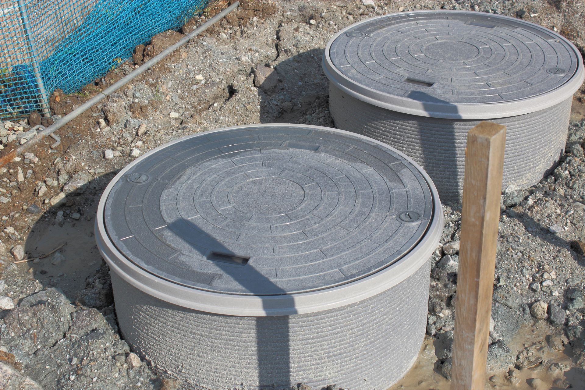Two round concrete utility access covers in a gravel yard beside a wooden stake and blue fence Two round concrete utility access covers in a gravel yard beside a wooden stake and blue fence