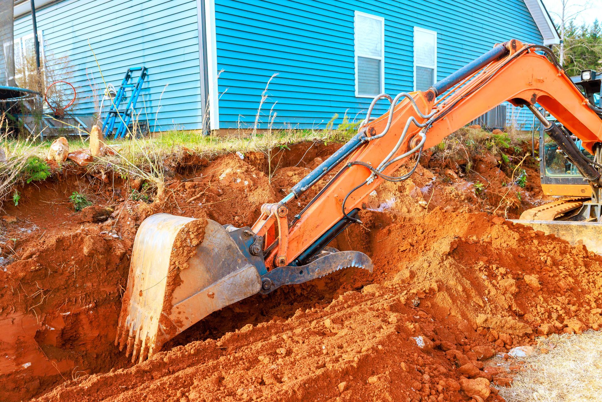 Orange excavator digging red dirt beside a blue house during construction Orange excavator digging red dirt beside a blue house during construction
