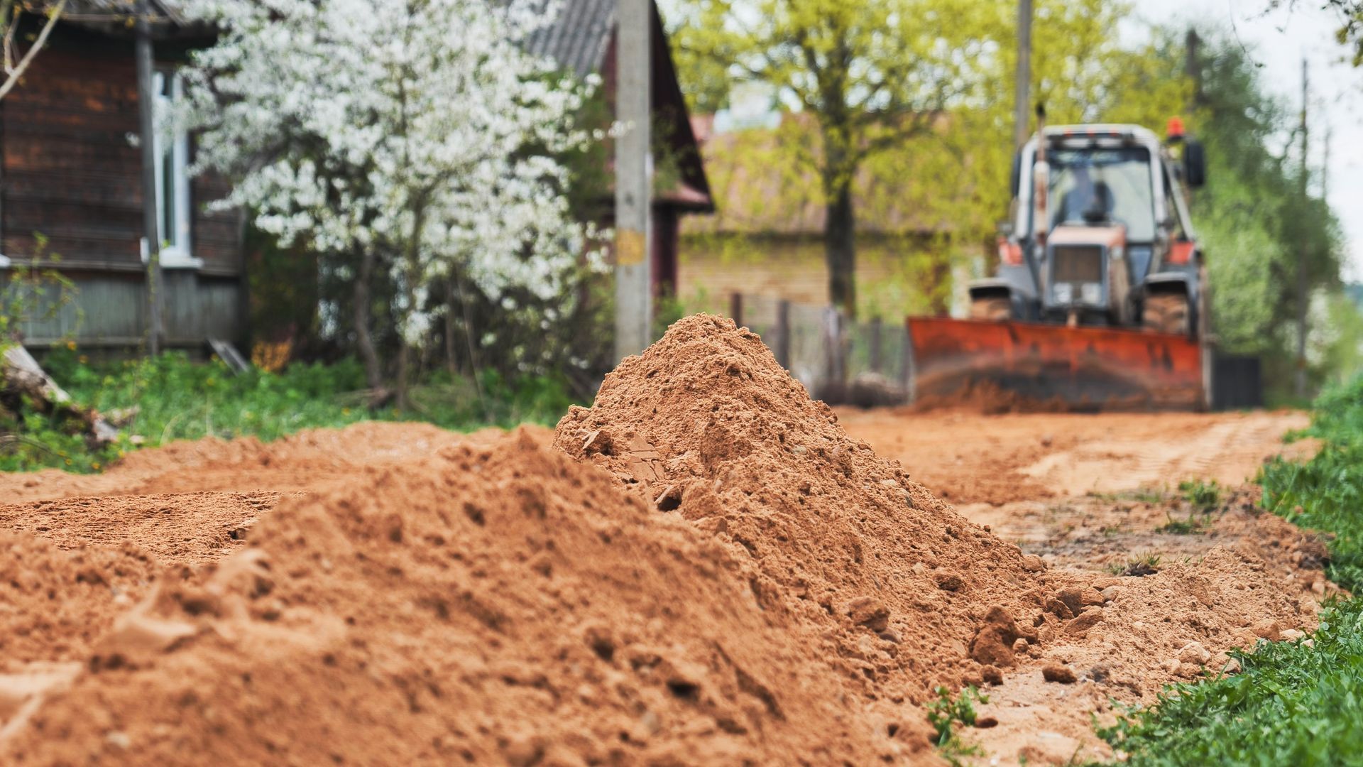 Fresh soil piled along a path while a backhoe works near houses and blooming trees Fresh soil piled along a path while a backhoe works near houses and blooming trees