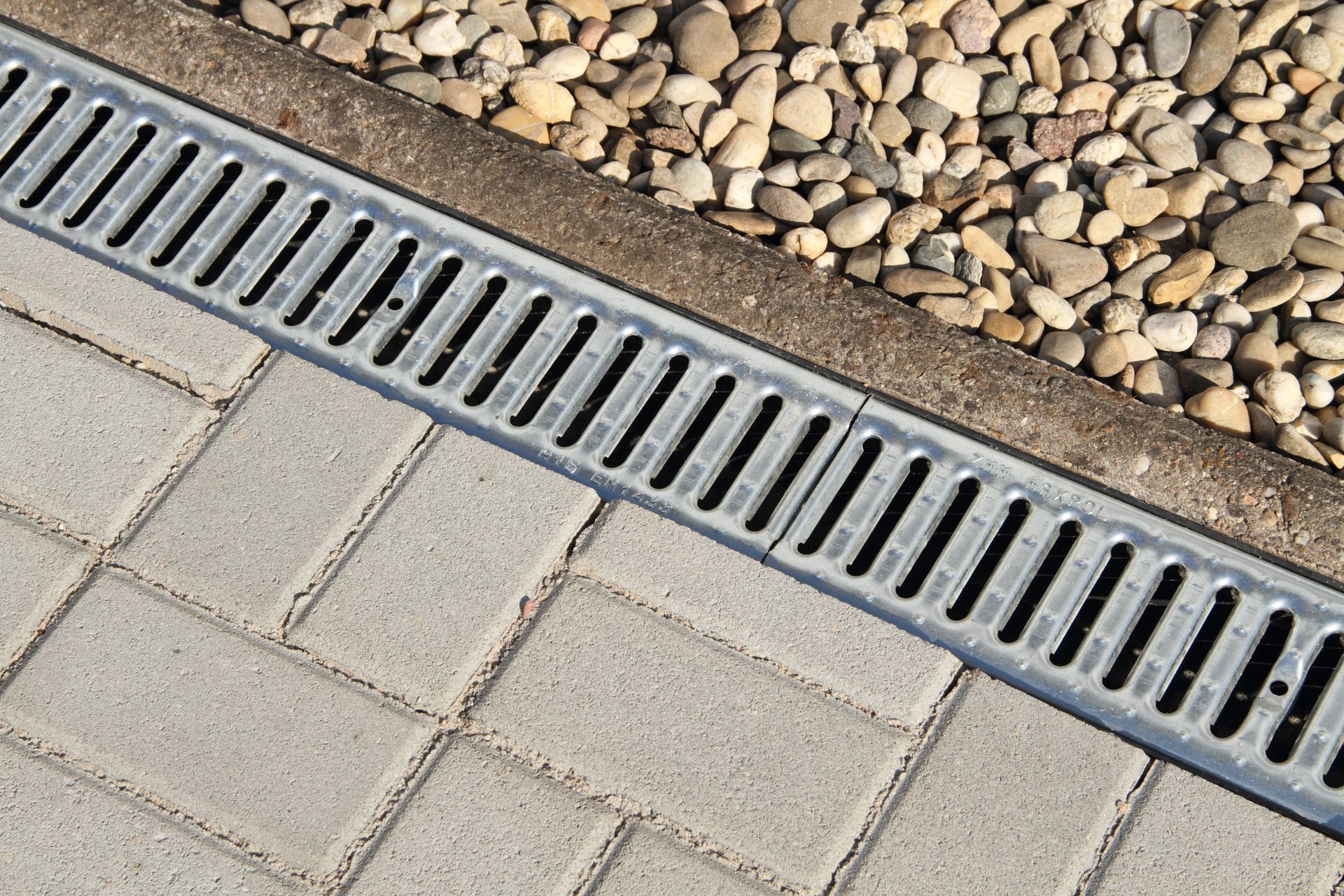 Drain grate between light gray paving stones and a pebble bed border Drain grate between light gray paving stones and a pebble bed border