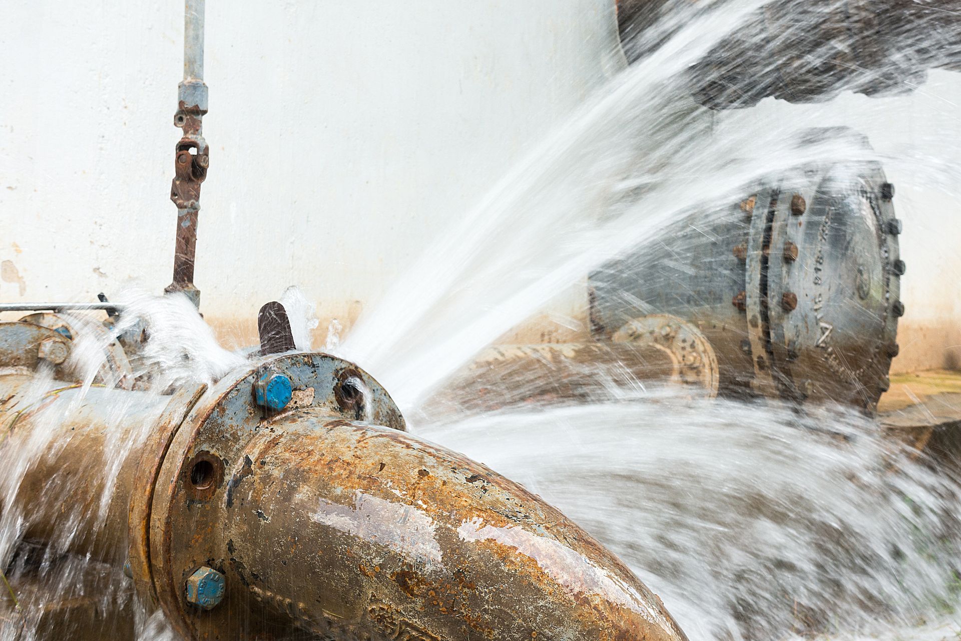 Water spraying from a rusted industrial pipe or valve in a metal facility, with a large tank in the background