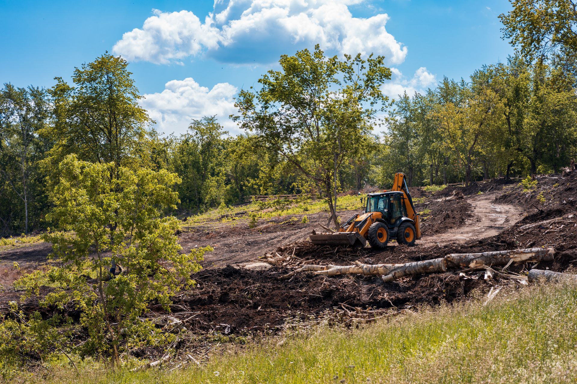 Backhoe clearing dirt on a wooded hillside under a blue sky with clouds Backhoe clearing dirt on a wooded hillside under a blue sky with clouds