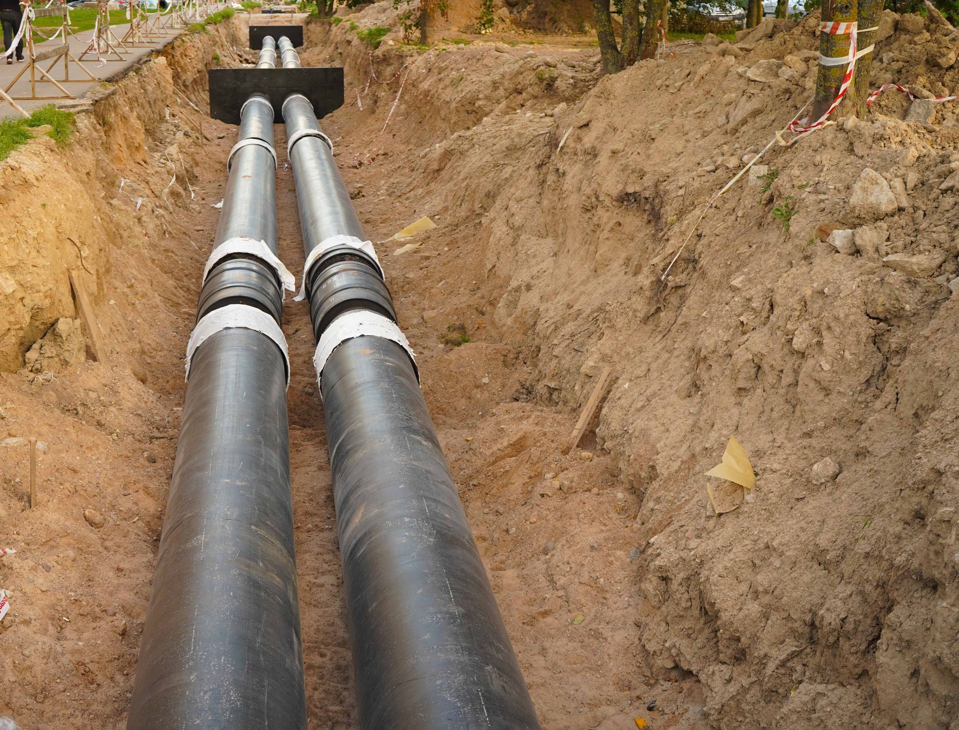 Two large black pipeline sections run through a dirt trench toward a culvert opening. Two large black pipeline sections run through a dirt trench toward a culvert opening.