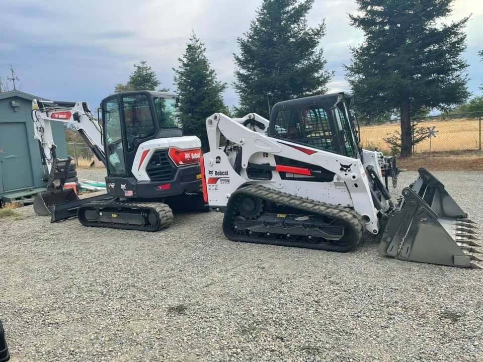 Two compact excavators, one white and one red, parked on gravel with evergreen trees in back Two compact excavators, one white and one red, parked on gravel with evergreen trees in back