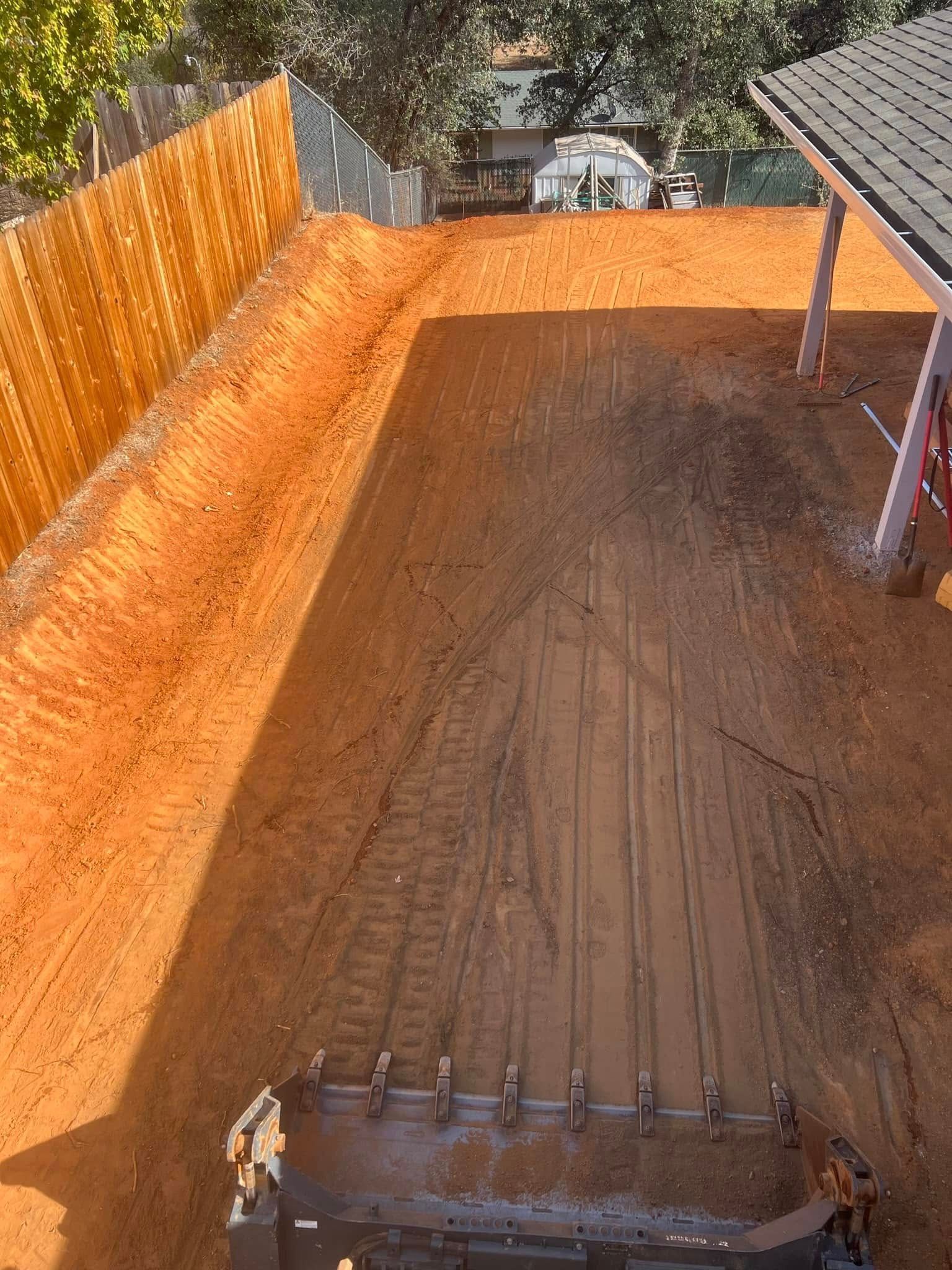 Backyard dirt excavation with orange soil, wooden fence, and a metal post frame on the right. Backyard dirt excavation with orange soil, wooden fence, and a metal post frame on the right.