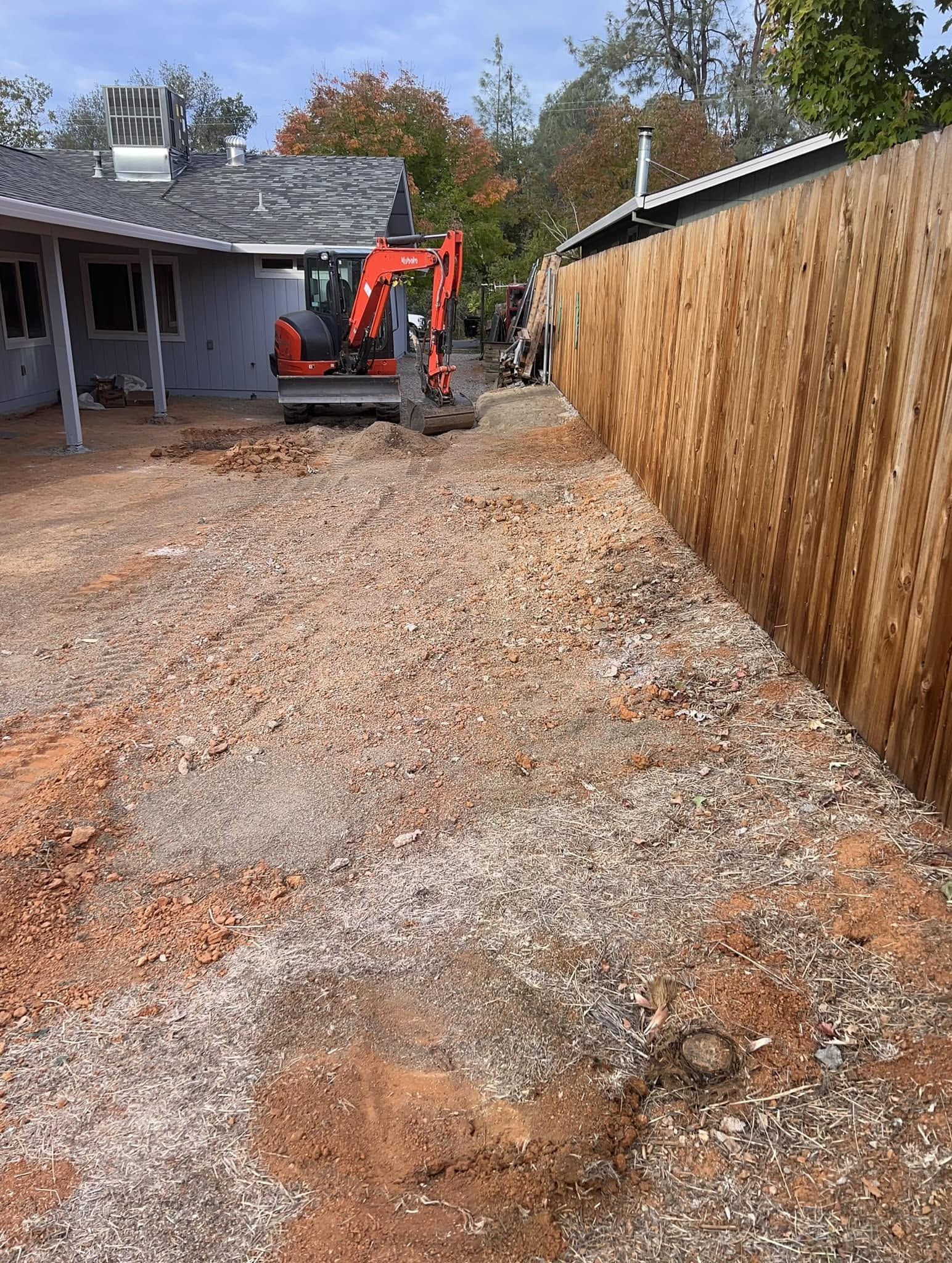 Orange excavator beside a house, grading a gravel driveway next to a tall wooden fence. Orange excavator beside a house, grading a gravel driveway next to a tall wooden fence.