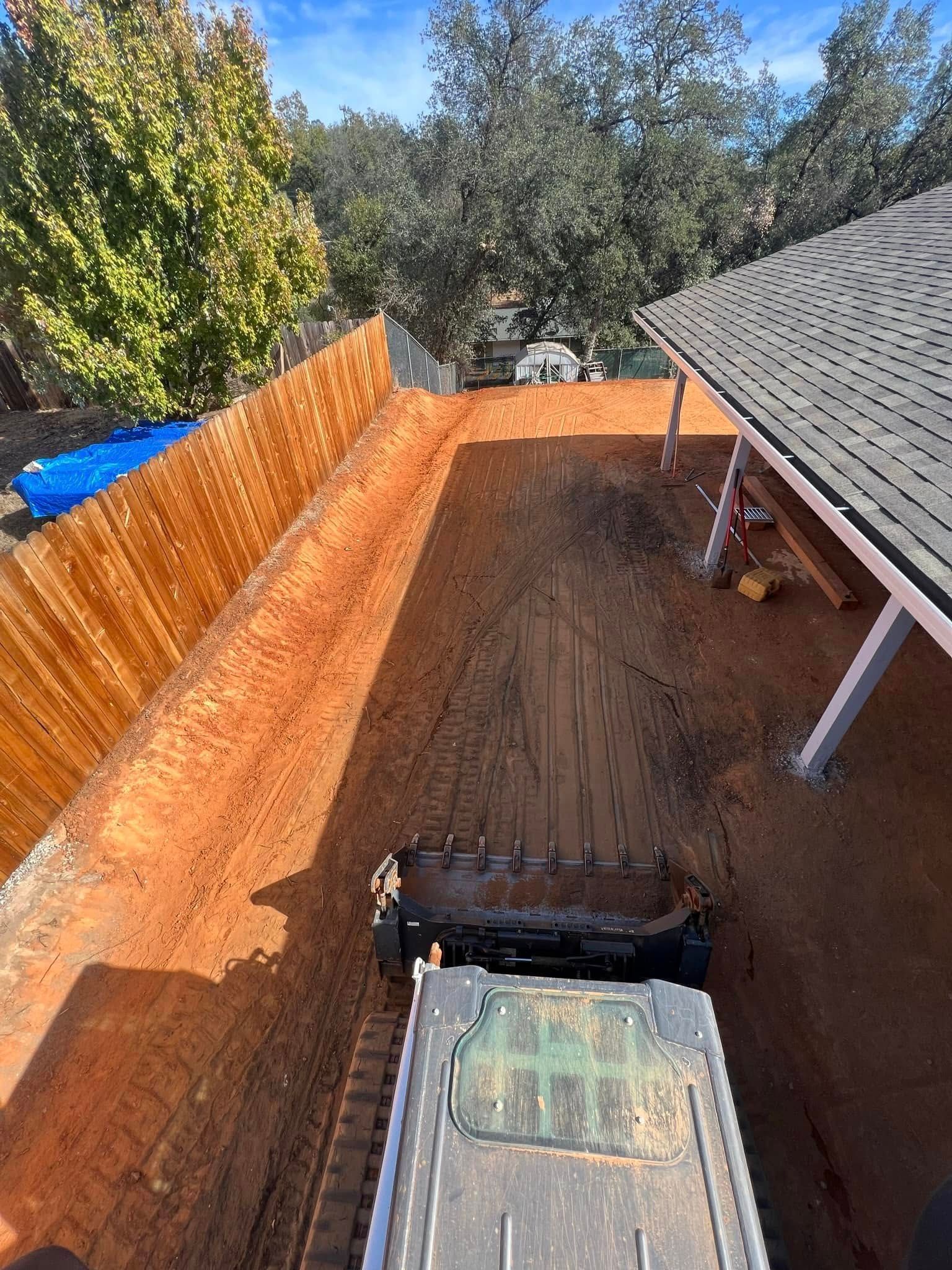 Long dirt trench beside a house with orange fencing, roof eave, and a front loader bucket in the foreground Long dirt trench beside a house with orange fencing, roof eave, and a front loader bucket in the foreground