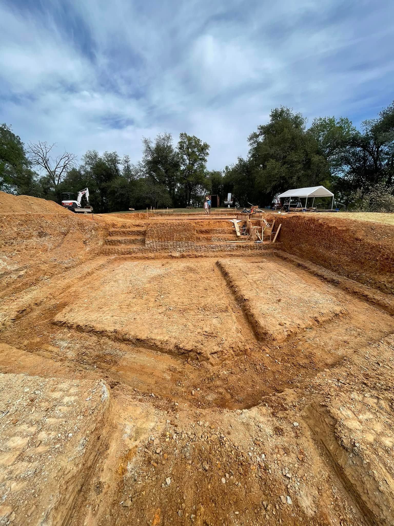 Large excavated dirt pit at a construction site under a cloudy sky, with workers and vehicles on the rim Large excavated dirt pit at a construction site under a cloudy sky, with workers and vehicles on the rim