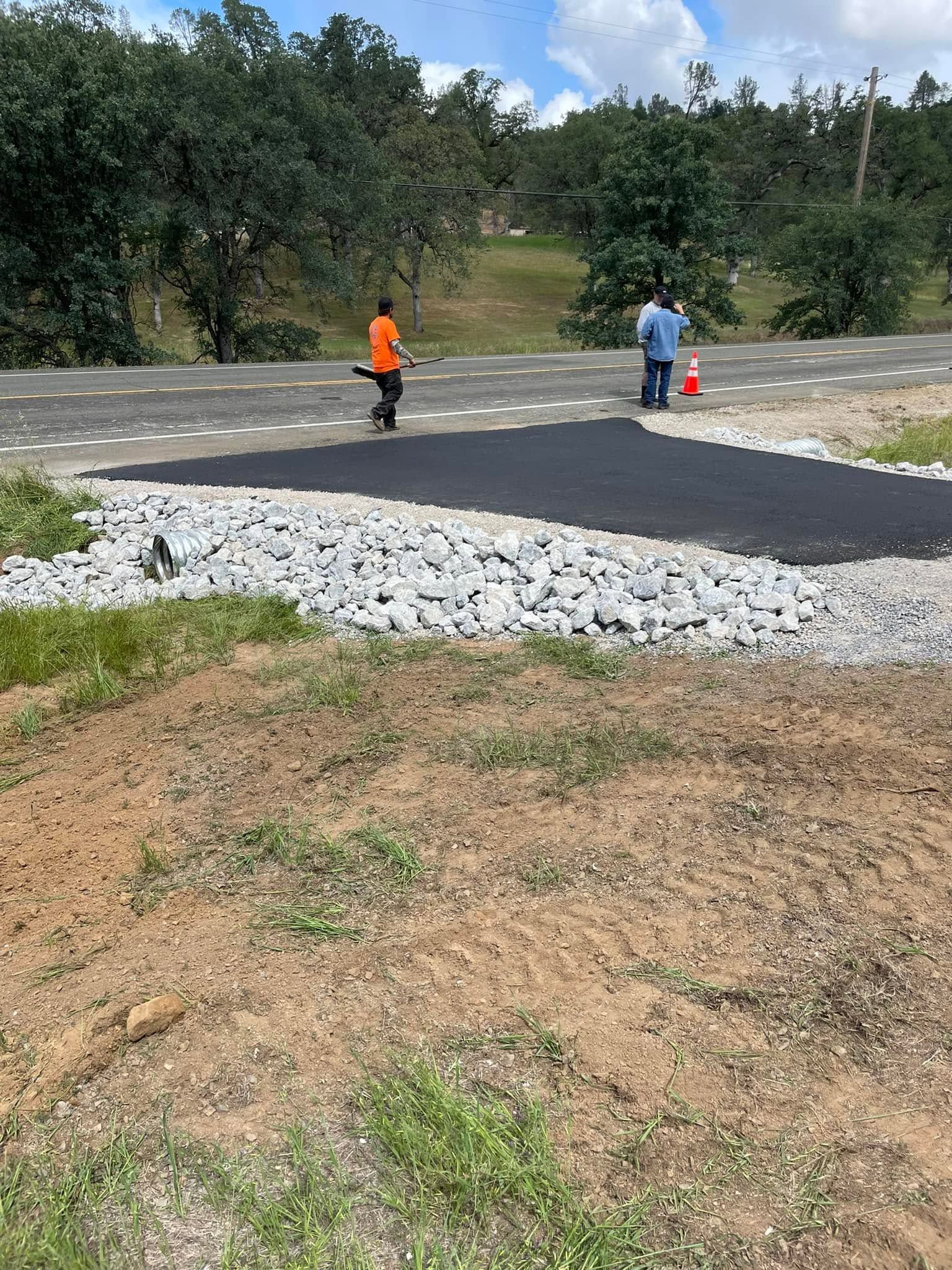 Workers paving a dark road section in a grassy rural area, with trees in the background. Workers paving a dark road section in a grassy rural area, with trees in the background.