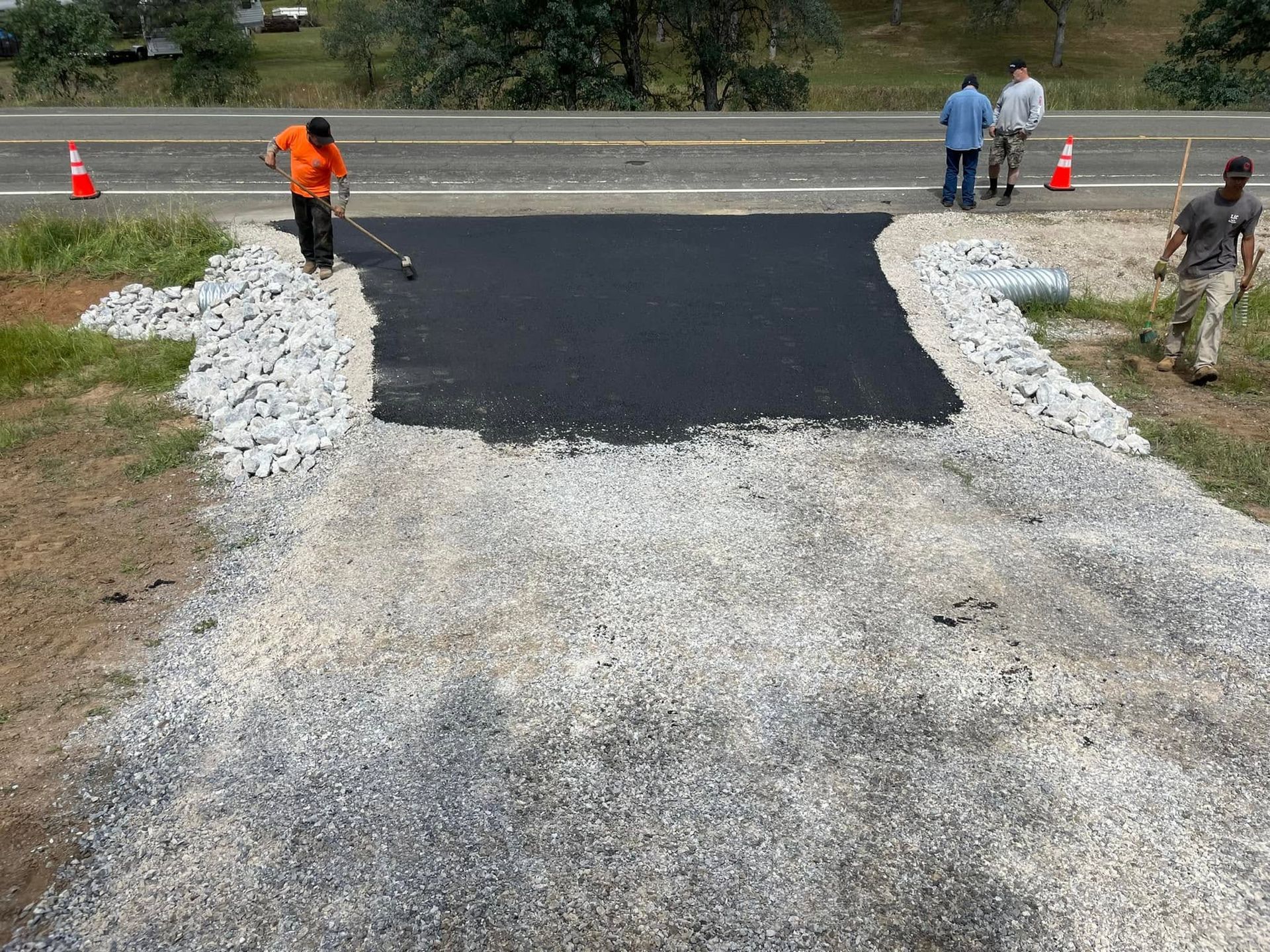 Workers paving a dark asphalt strip over a gravel road near a roadside, with cones and onlookers nearby Workers paving a dark asphalt strip over a gravel road near a roadside, with cones and onlookers nearby