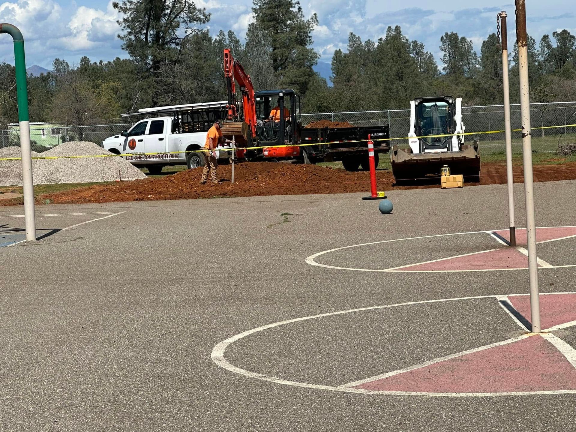 Construction vehicles and orange cones on a gravel court beside pink basketball circles. Construction vehicles and orange cones on a gravel court beside pink basketball circles.