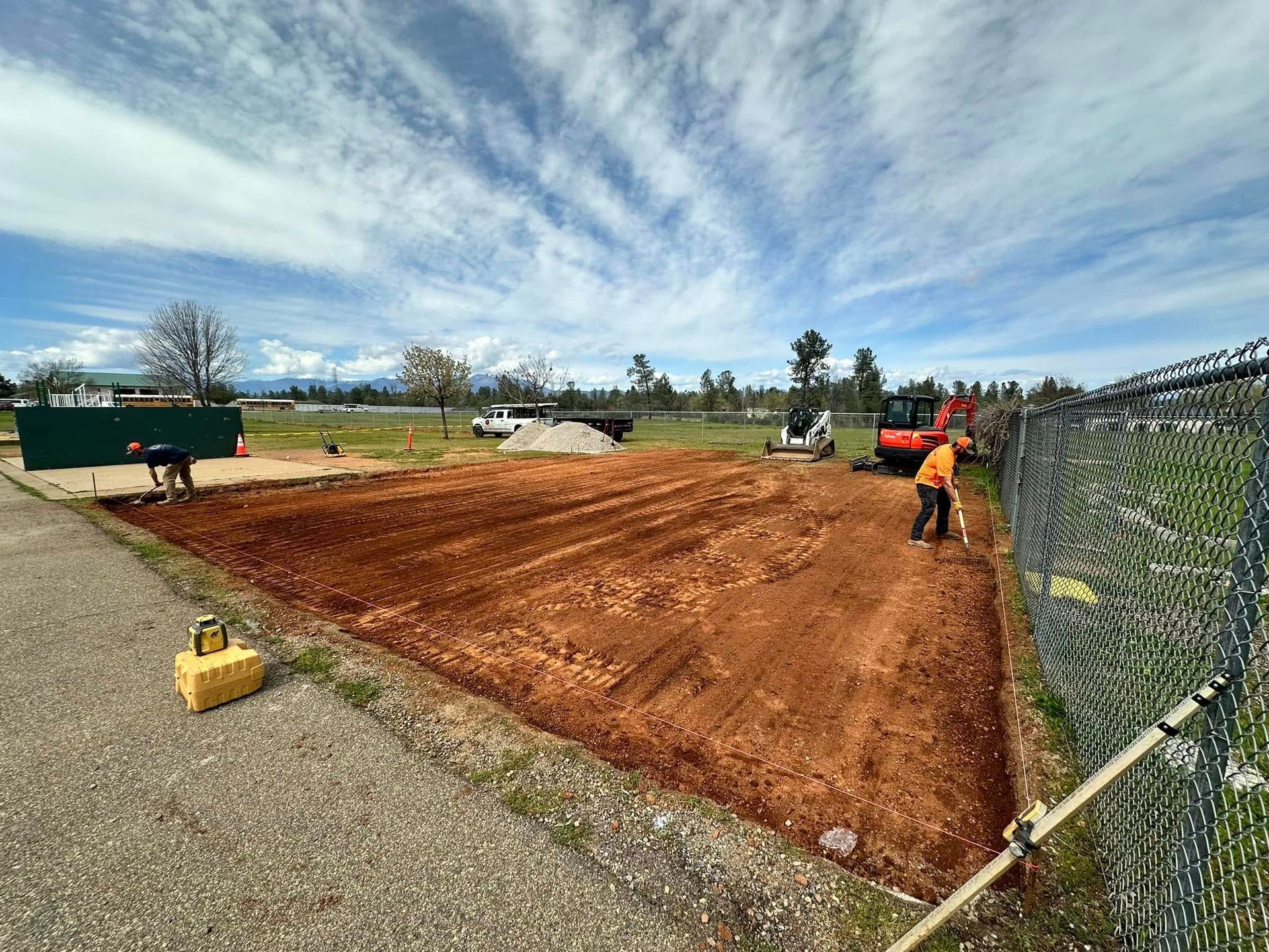 Construction site with red dirt, workers, heavy equipment, and a chain-link fence under a cloudy sky Construction site with red dirt, workers, heavy equipment, and a chain-link fence under a cloudy sky