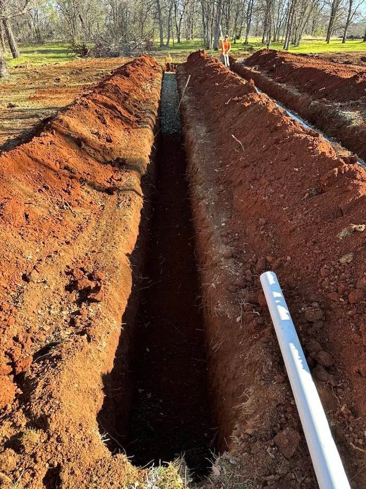 Red dirt trench dug through a wooded area, with a white pipe running along the right side. Red dirt trench dug through a wooded area, with a white pipe running along the right side.