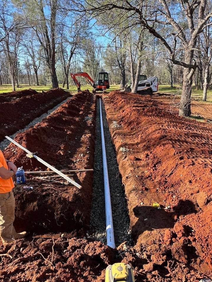 Workers install a long black pipe in a muddy trench beside an orange excavator in a wooded area Workers install a long black pipe in a muddy trench beside an orange excavator in a wooded area