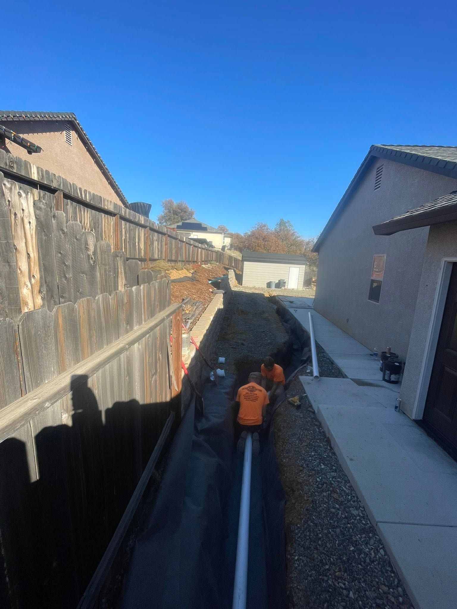Person in orange jacket inspecting a narrow trench between a wooden fence and a house on a sunny day Person in orange jacket inspecting a narrow trench between a wooden fence and a house on a sunny day