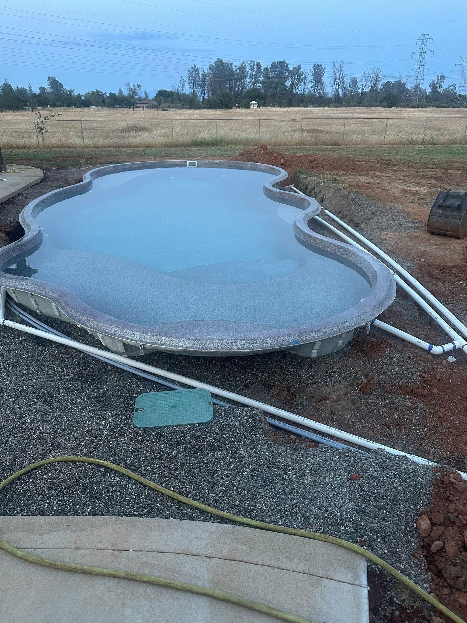 Kidney-shaped backyard pool under construction, filled with water and surrounded by gravel and pipes. Kidney-shaped backyard pool under construction, filled with water and surrounded by gravel and pipes.