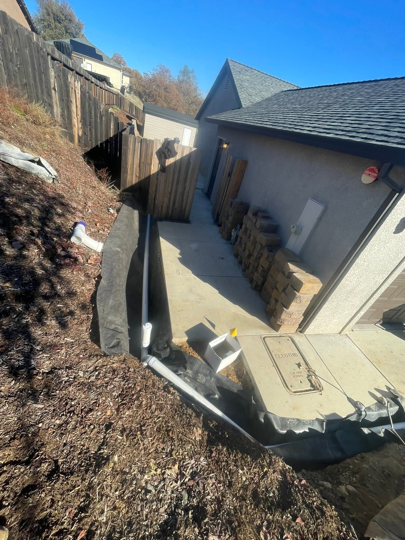 Backyard patio with a house wall, retaining wall, and narrow concrete walkway beside a hillside. Backyard patio with a house wall, retaining wall, and narrow concrete walkway beside a hillside.
