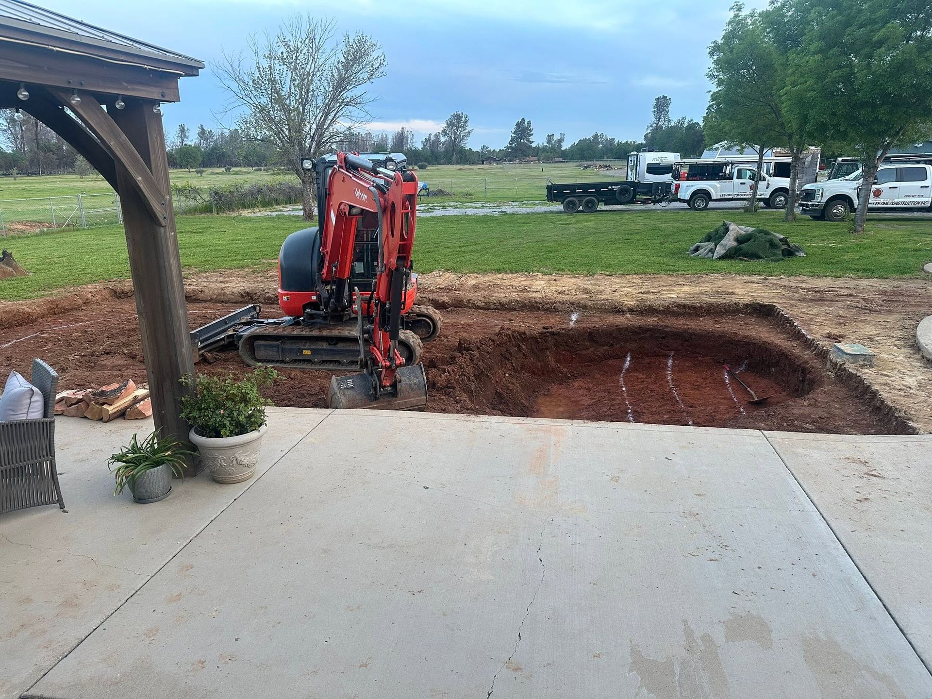 Red excavator digging a large hole beside a concrete slab in a rural yard. Red excavator digging a large hole beside a concrete slab in a rural yard.