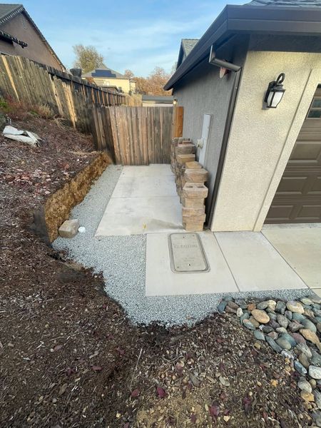 Narrow concrete side yard path beside a house, bordered by gravel, rocks, and a wooden fence. Narrow concrete side yard path beside a house, bordered by gravel, rocks, and a wooden fence.