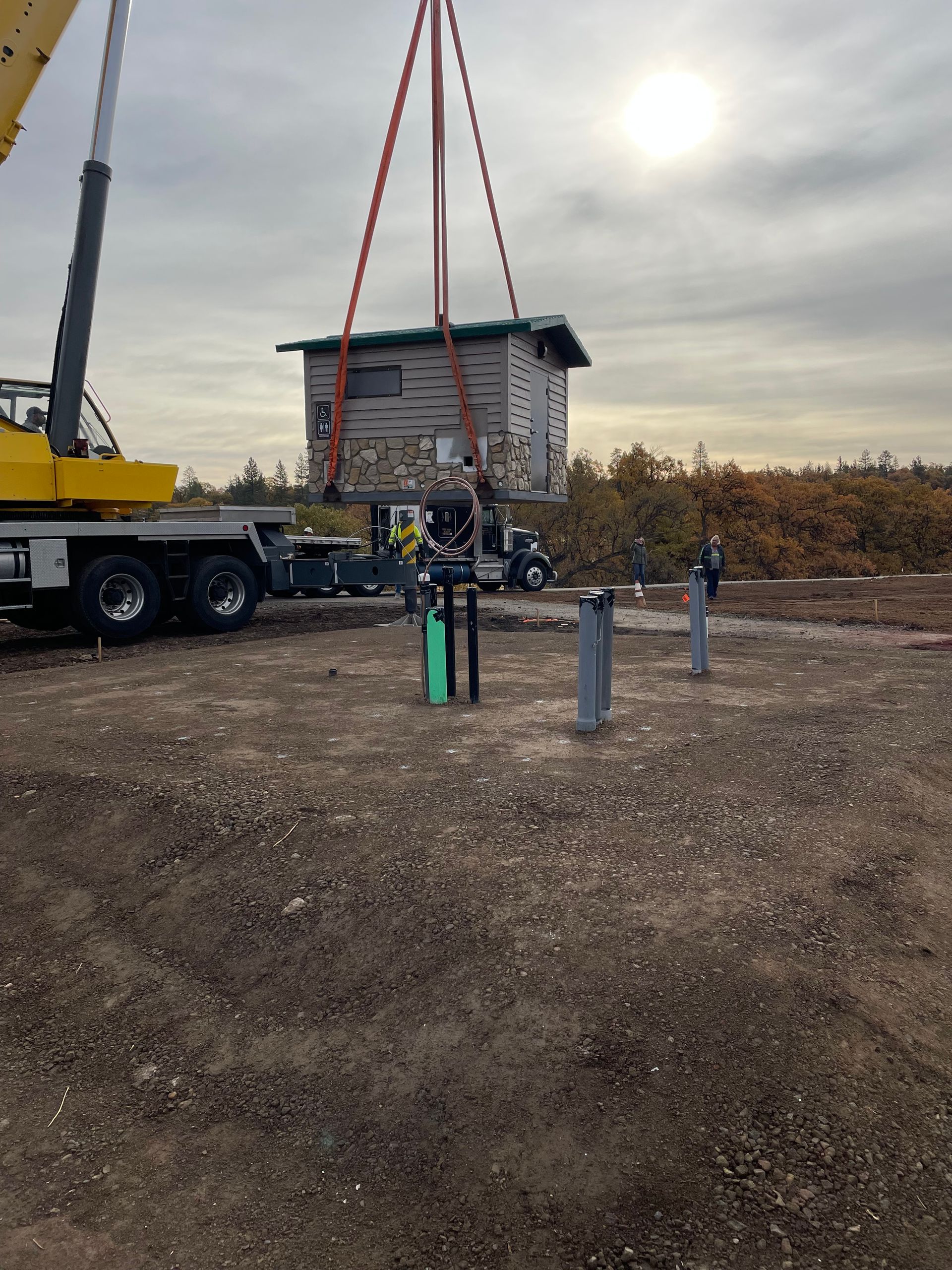 Crane lifting a small cabin beside a truck in a gravel lot at sunset
