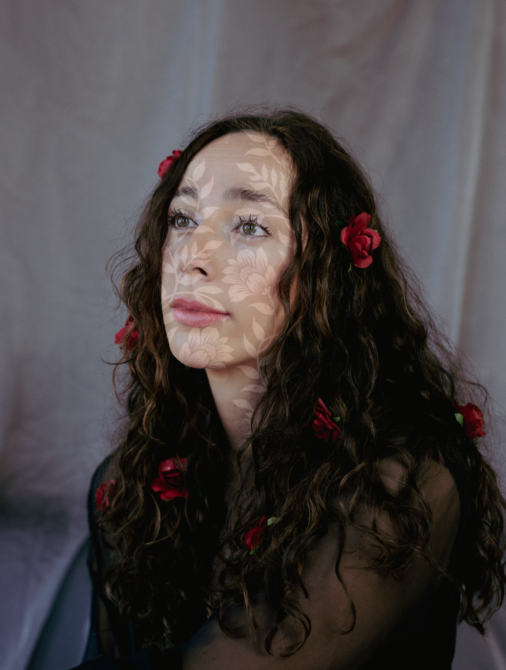 Persoon met lang donker krullend haar en rode bloemen, poserend tegen een grijze achtergrond.