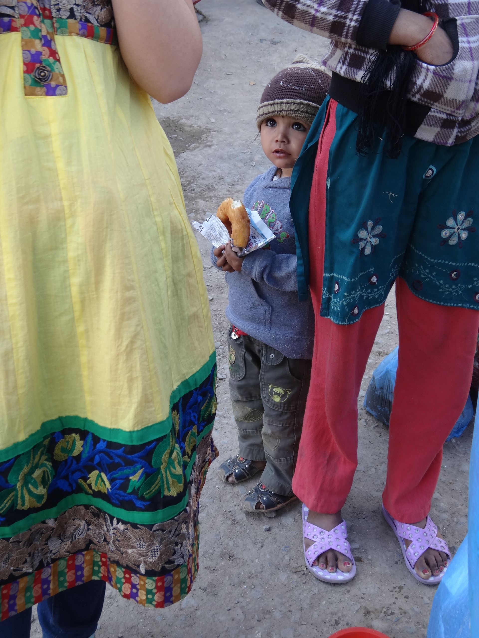 Een kind met een snack staat buiten naast volwassenen in kleurrijke kleding.
