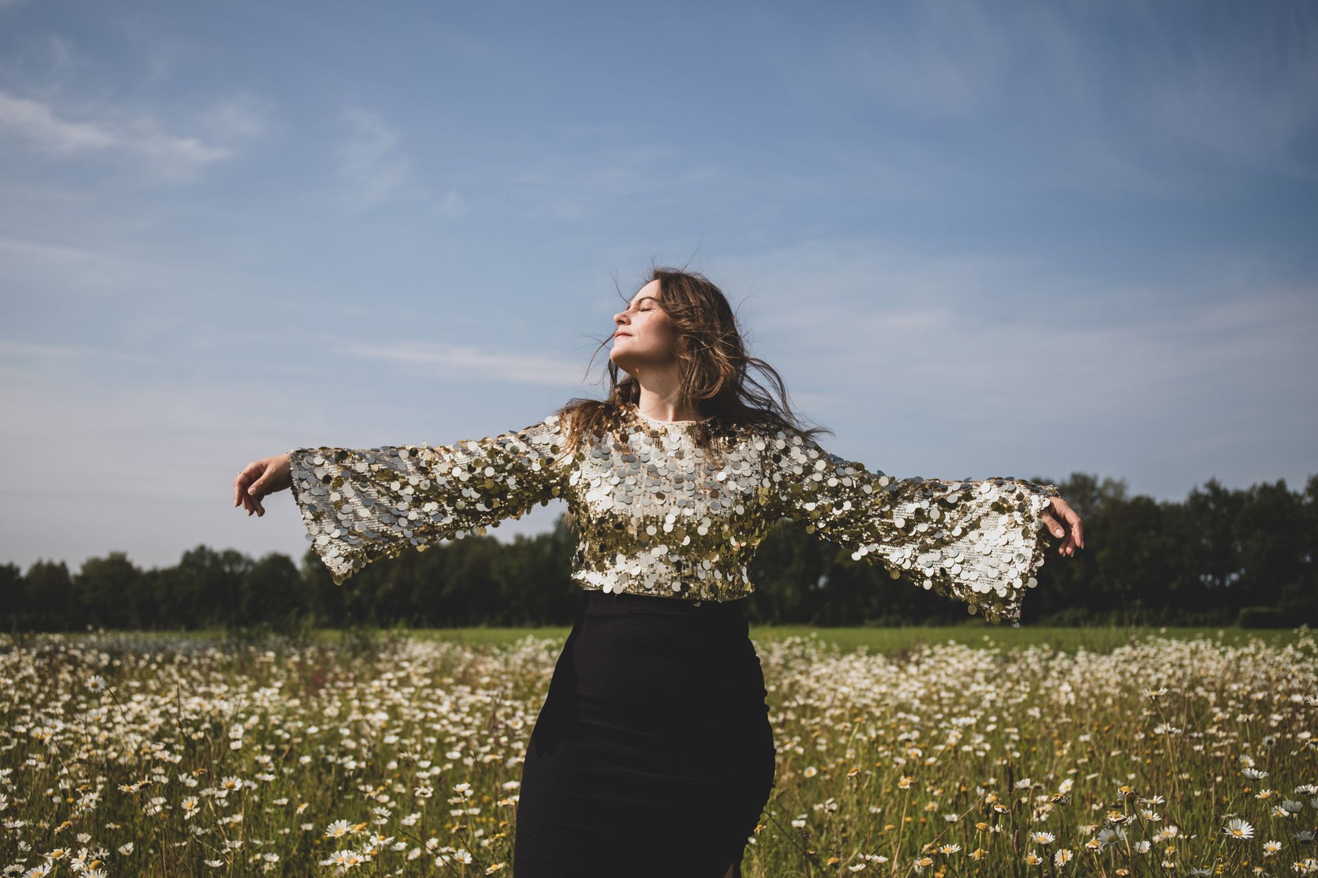 Vrouw in gouden top en een zwarte rok staat in een veld met wilde bloemen, met de armen uitgestrekt onder een blauwe hemel.