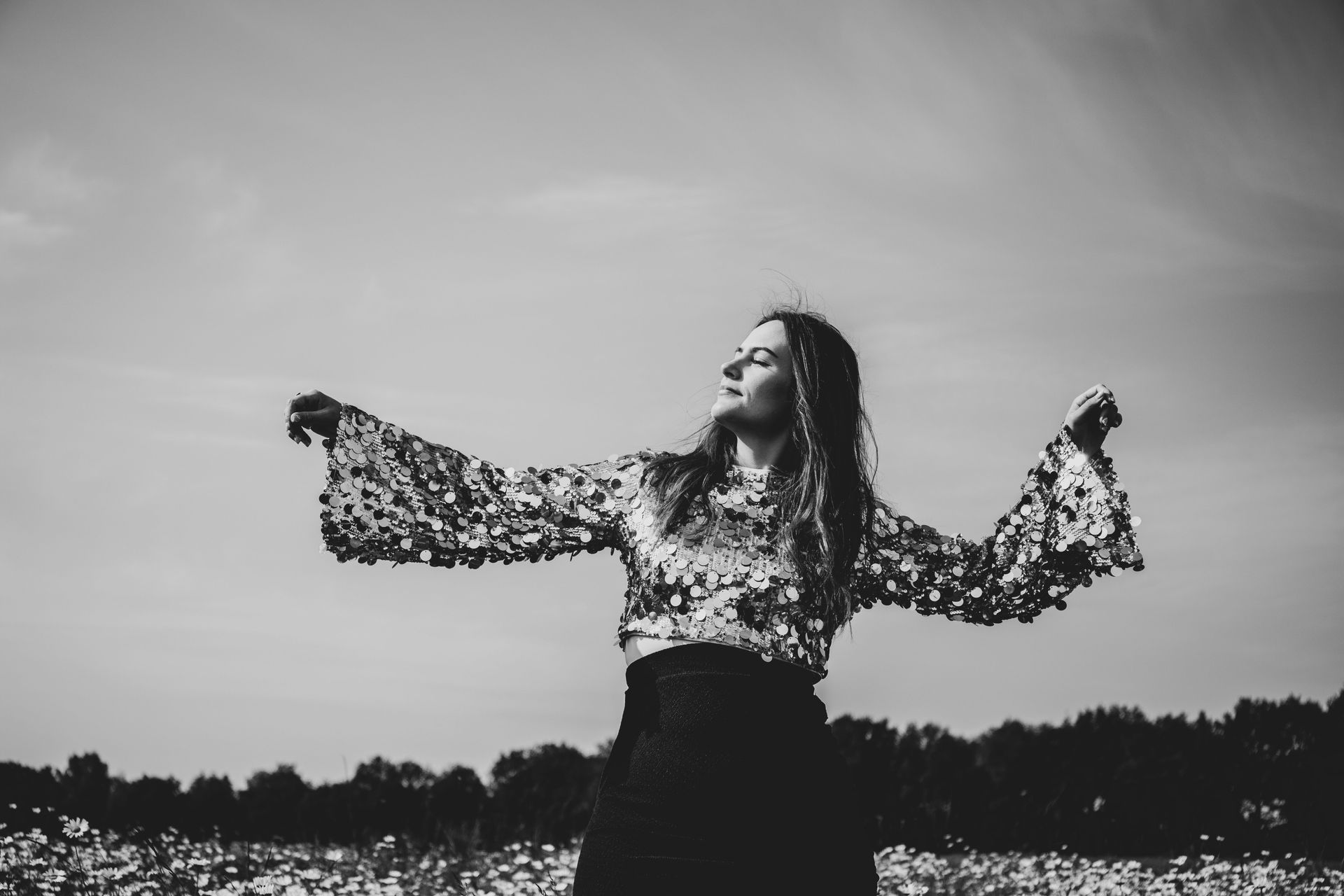 Een persoon in een blouse met een patroon staat in een veld met opgeheven armen onder een bewolkte hemel.
