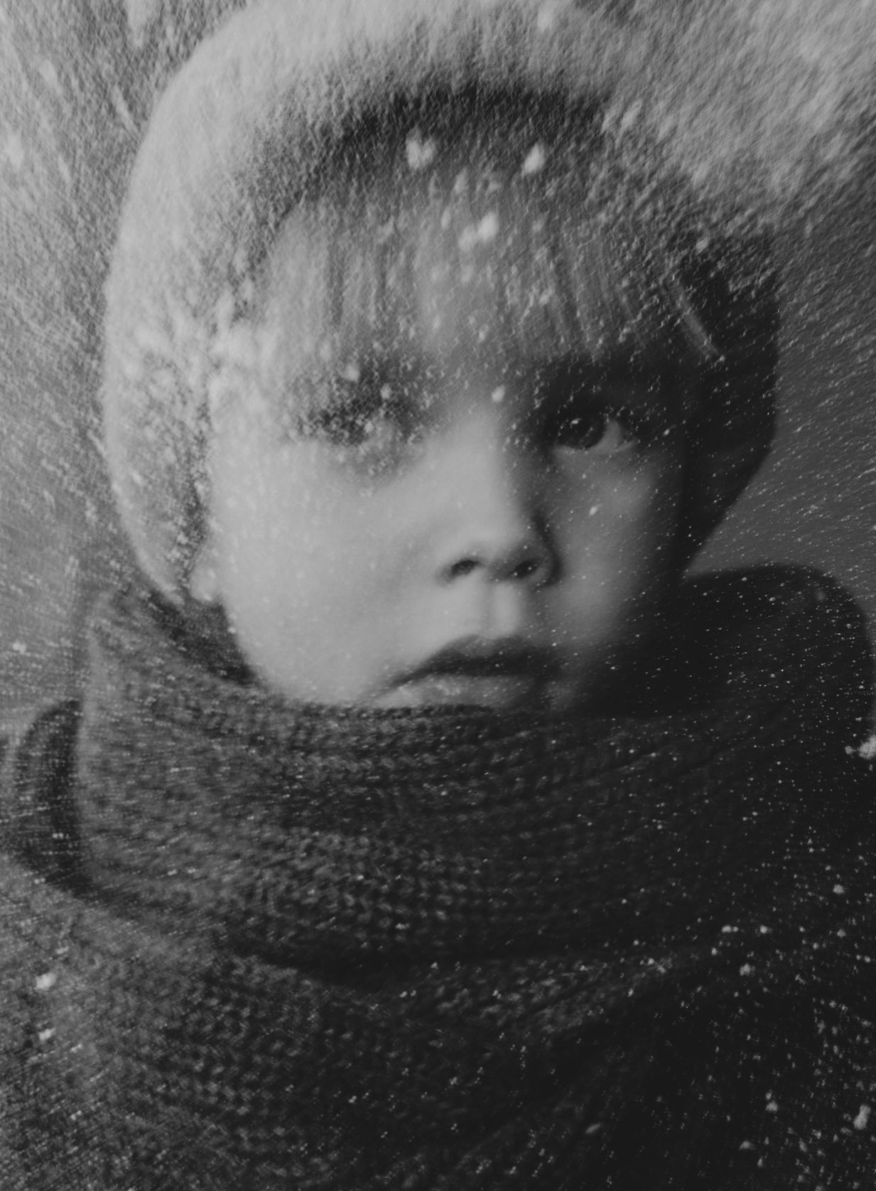 Kind, gewikkeld in een sjaal en een harige muts, met sneeuw op het gezicht, kijkt naar de camera.