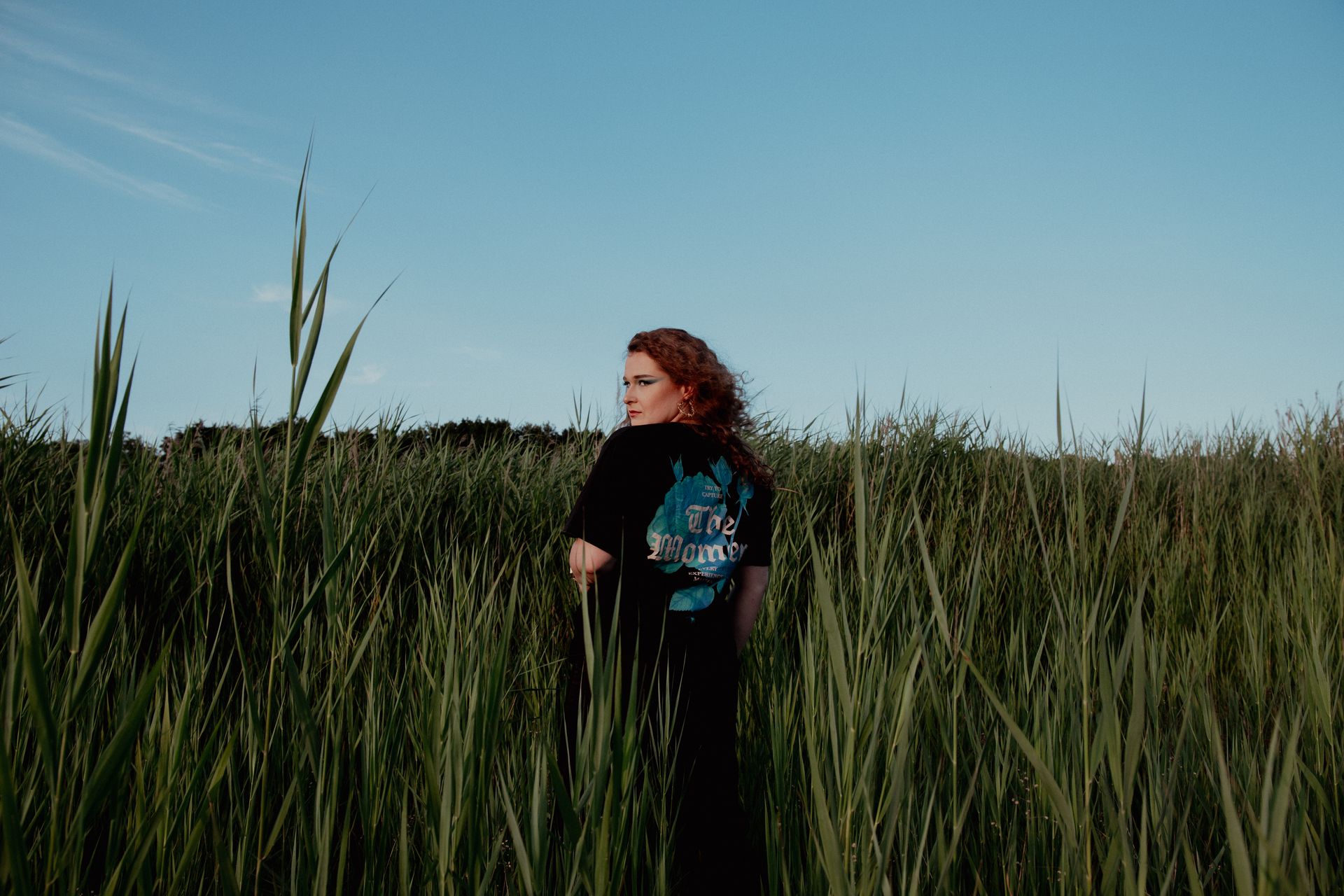Een persoon staat in hoog gras onder een blauwe hemel, gekleed in een zwart shirt met een kleurrijke afbeelding.