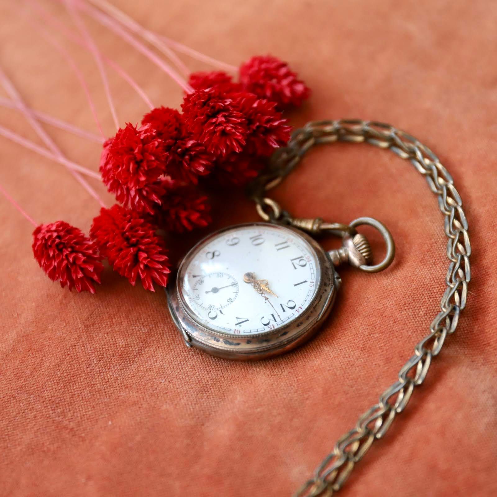 Vintage zakhorloge met zilveren ketting en rode pomponkwastjes op een perzikkleurige ondergrond.