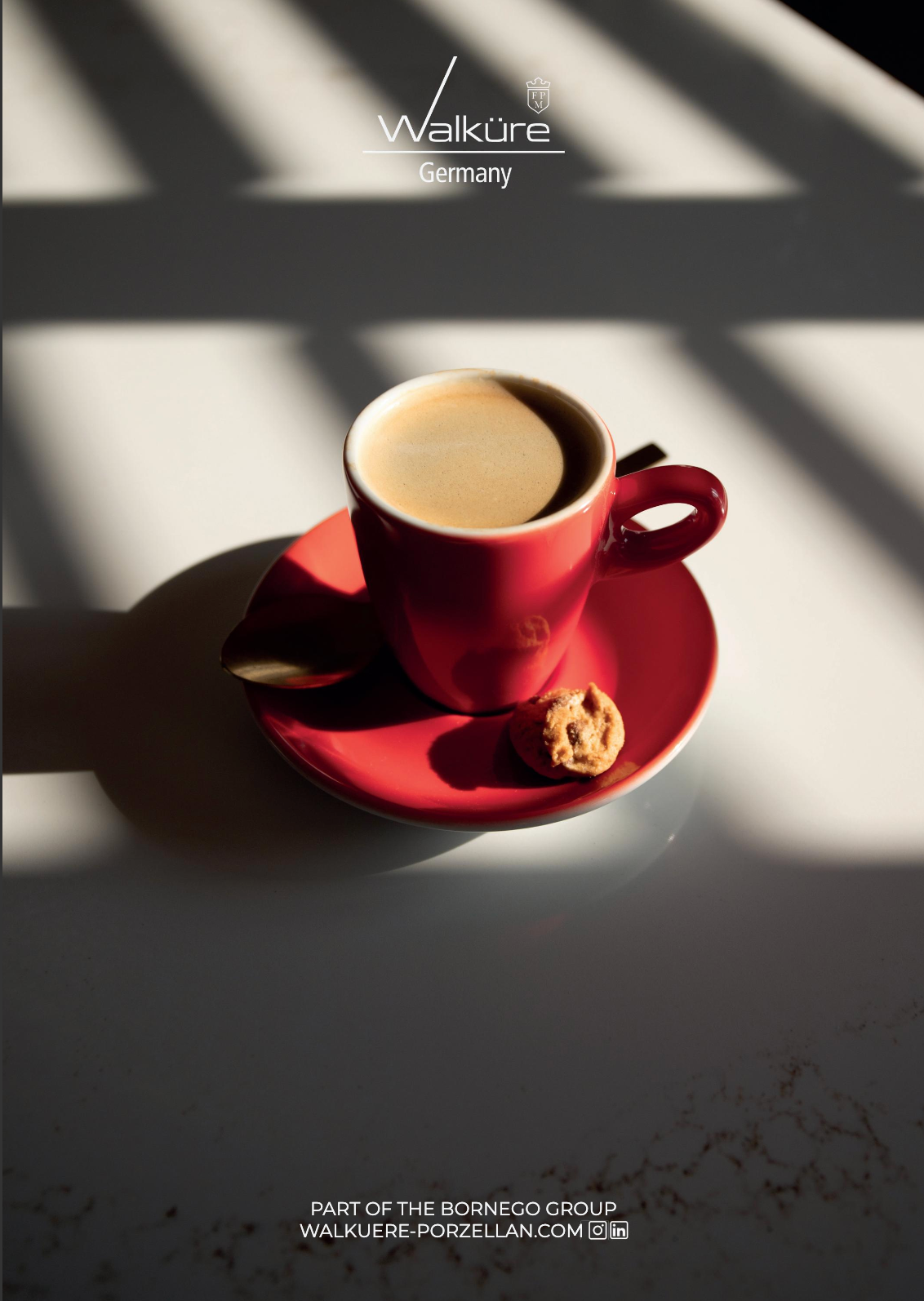Een rode kop koffie op een schoteltje met een koekje, verlicht door de schaduwen van het raam op een witte tafel.