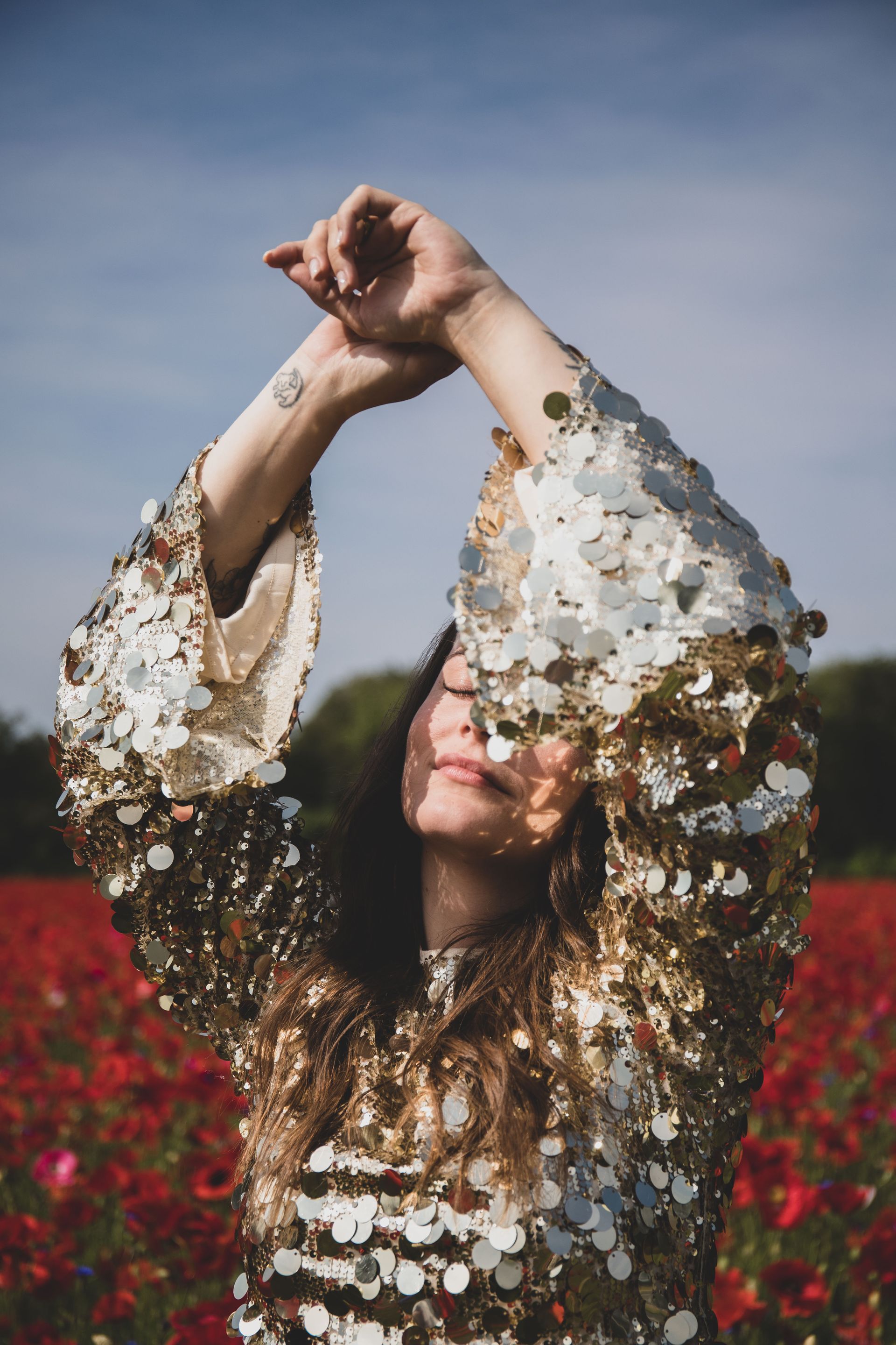Vrouw in een gouden paillettenjurk poseert met opgeheven armen in een veld met rode bloemen onder een blauwe hemel.