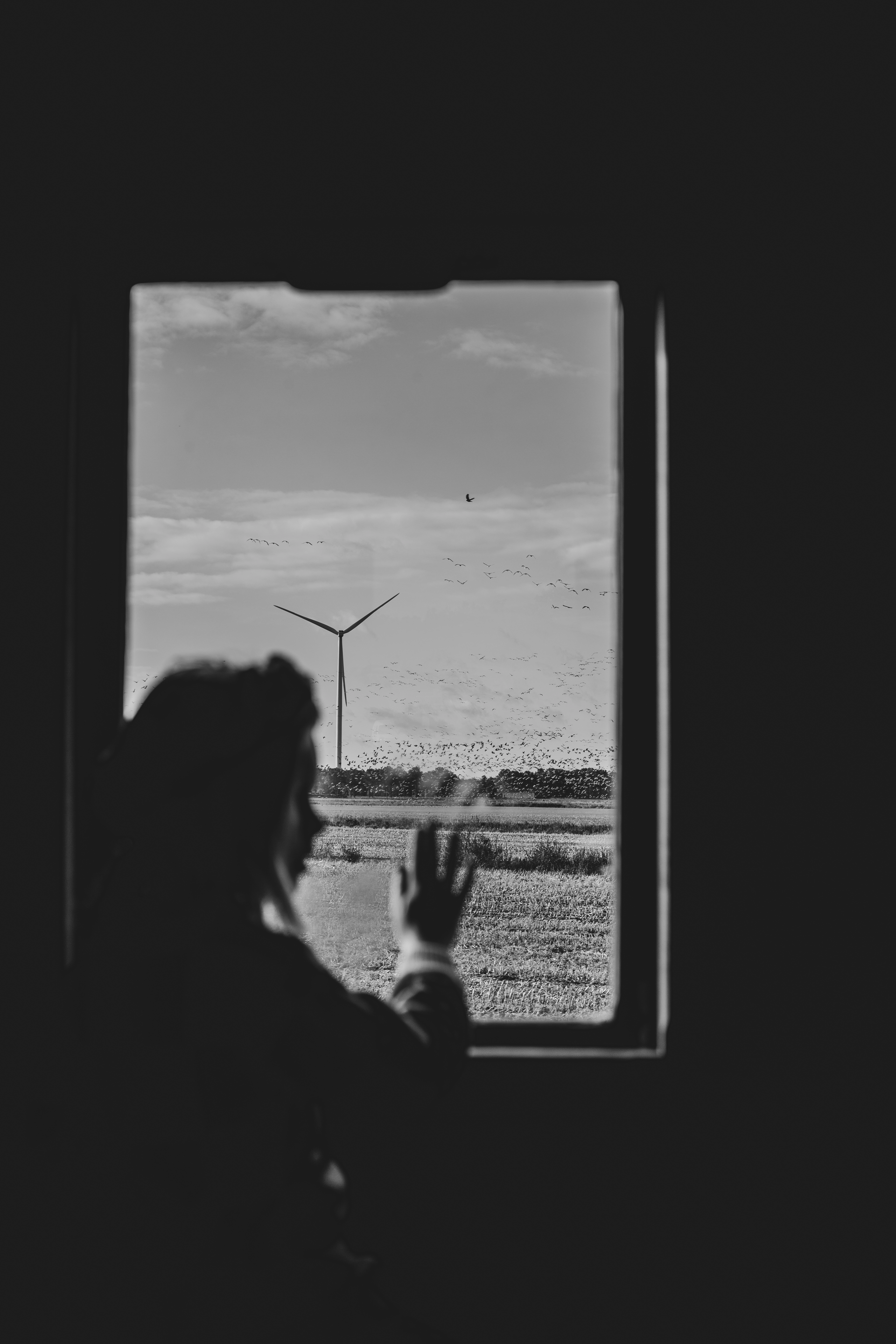 Silhouet van een persoon die vanuit een donkere kamer door een raam naar een bewolkte buitenomgeving zwaait.