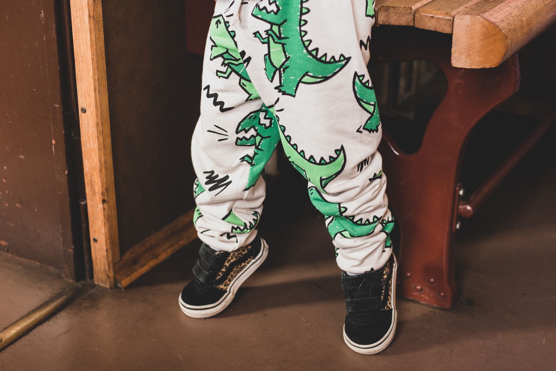 De benen van een kind in een witte broek met dinosaurusprint en zwarte sneakers staan binnen naast een houten stoel.