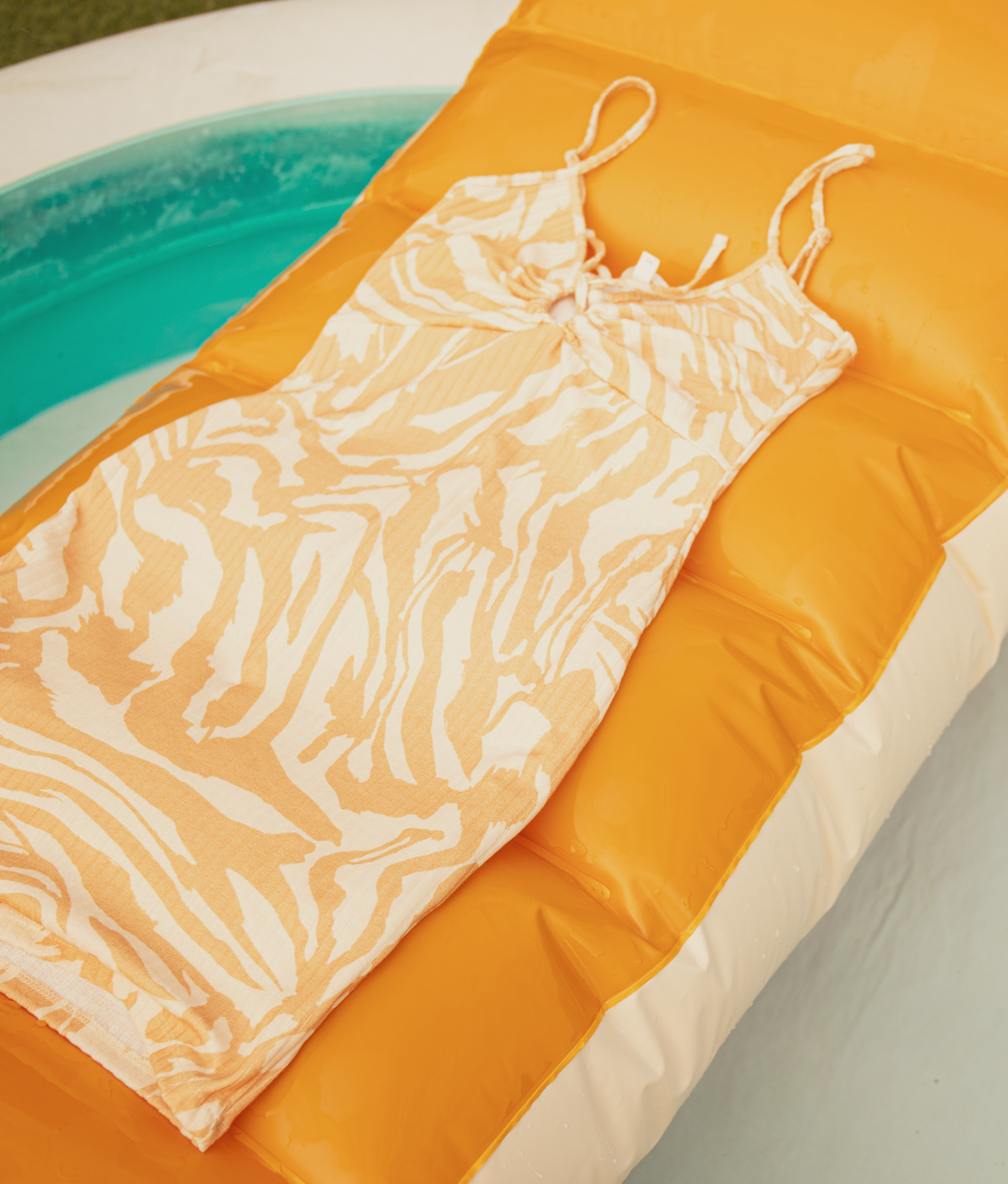 Een zomerjurk met print op een oranje opblaasbaar luchtbed naast een turquoise zwembad.