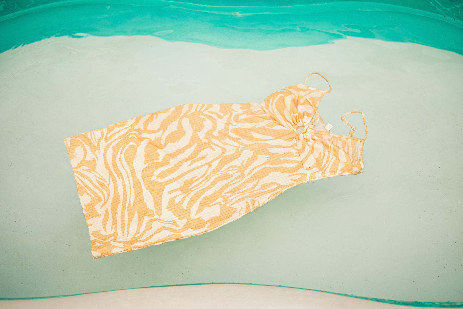 Een oranje jurk met patroon ligt plat op een lichtgekleurde ondergrond met turquoise water aan de boven- en onderkant.