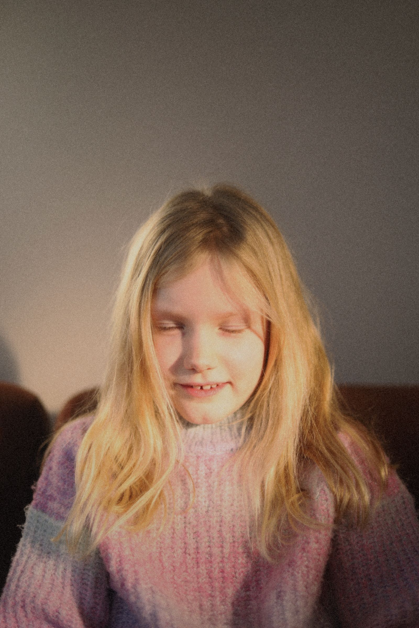 Jong persoon met lang blond haar, gekleed in een roze en paarse trui, kijkt naar beneden tegen een effen achtergrond.