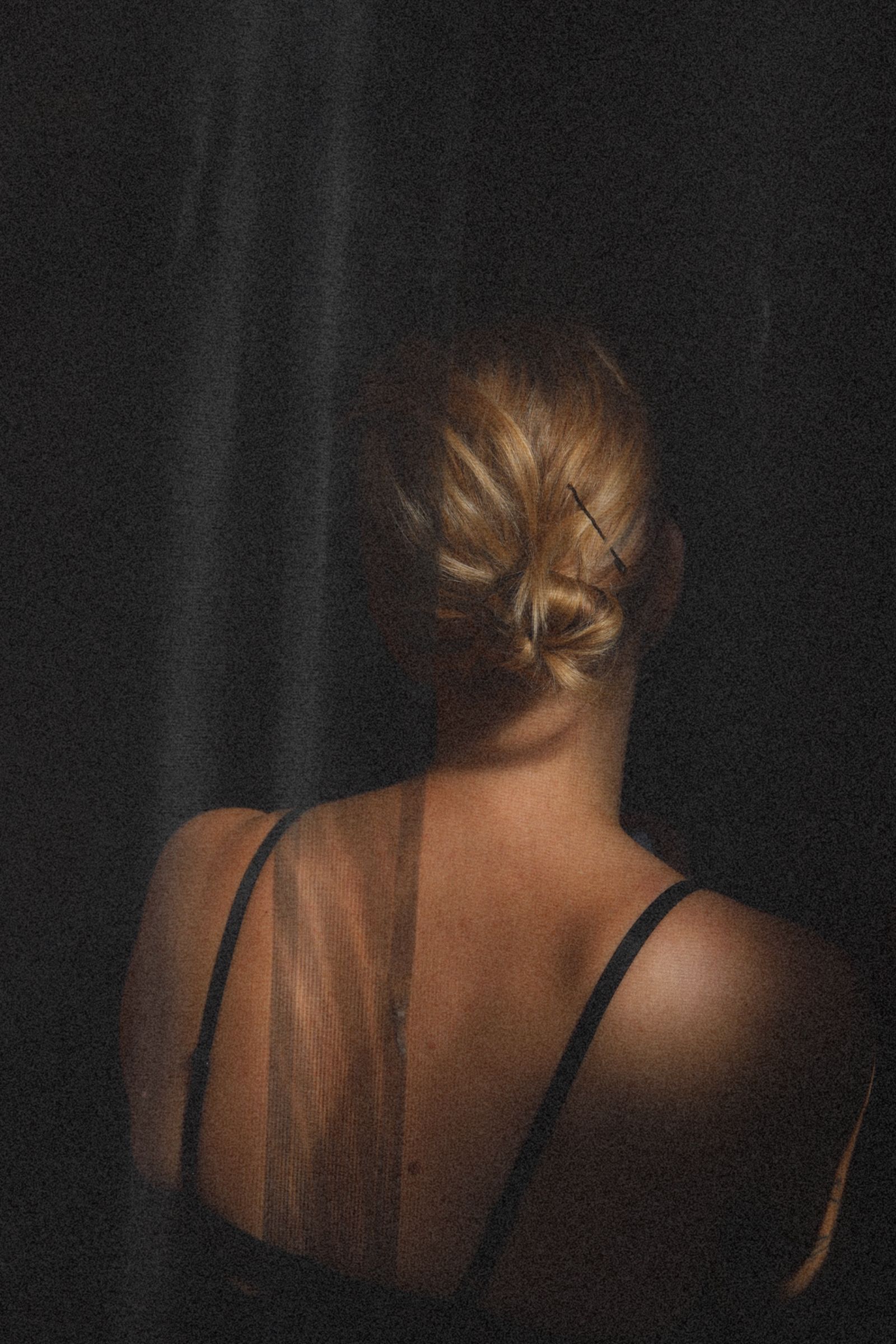 Achteraanzicht van een persoon met blond haar in een zwart hemdje tegen een donkere achtergrond.