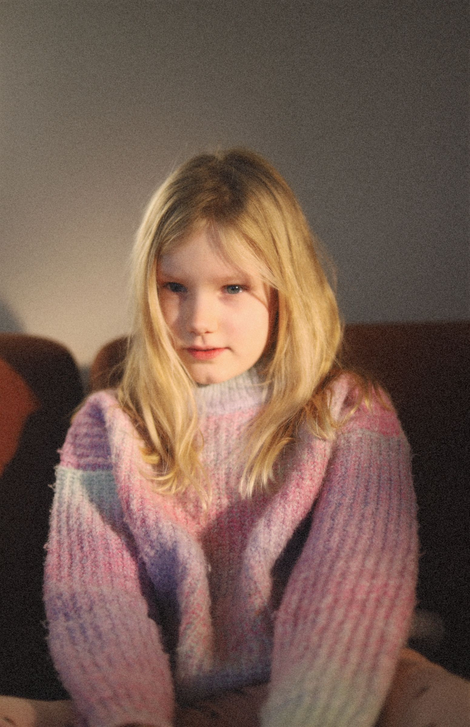 Blond kind in een roze en paarse trui zit op een bank en kijkt naar de camera.