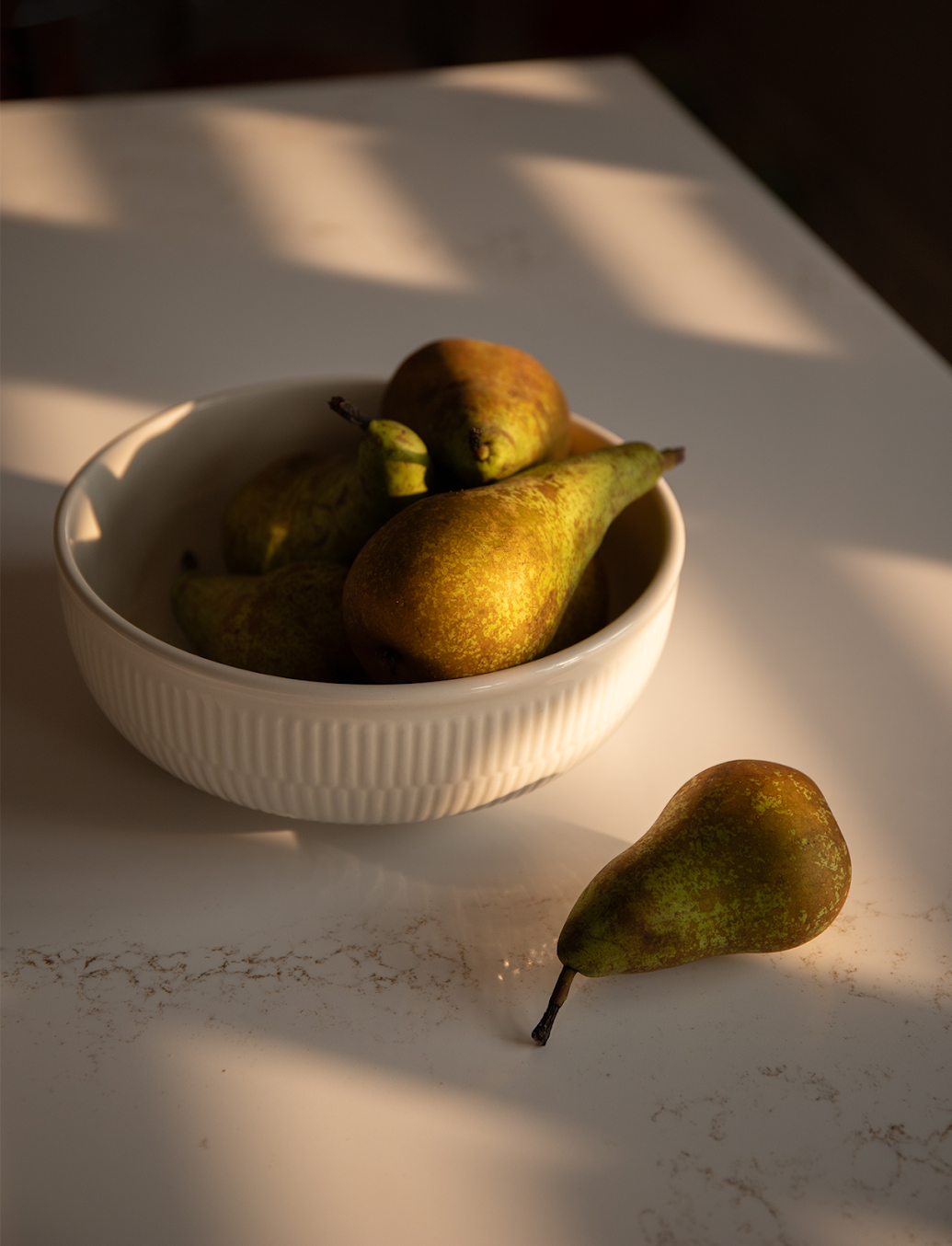 Een witte schaal met peren op een zonnige tafel, met één peer ernaast.