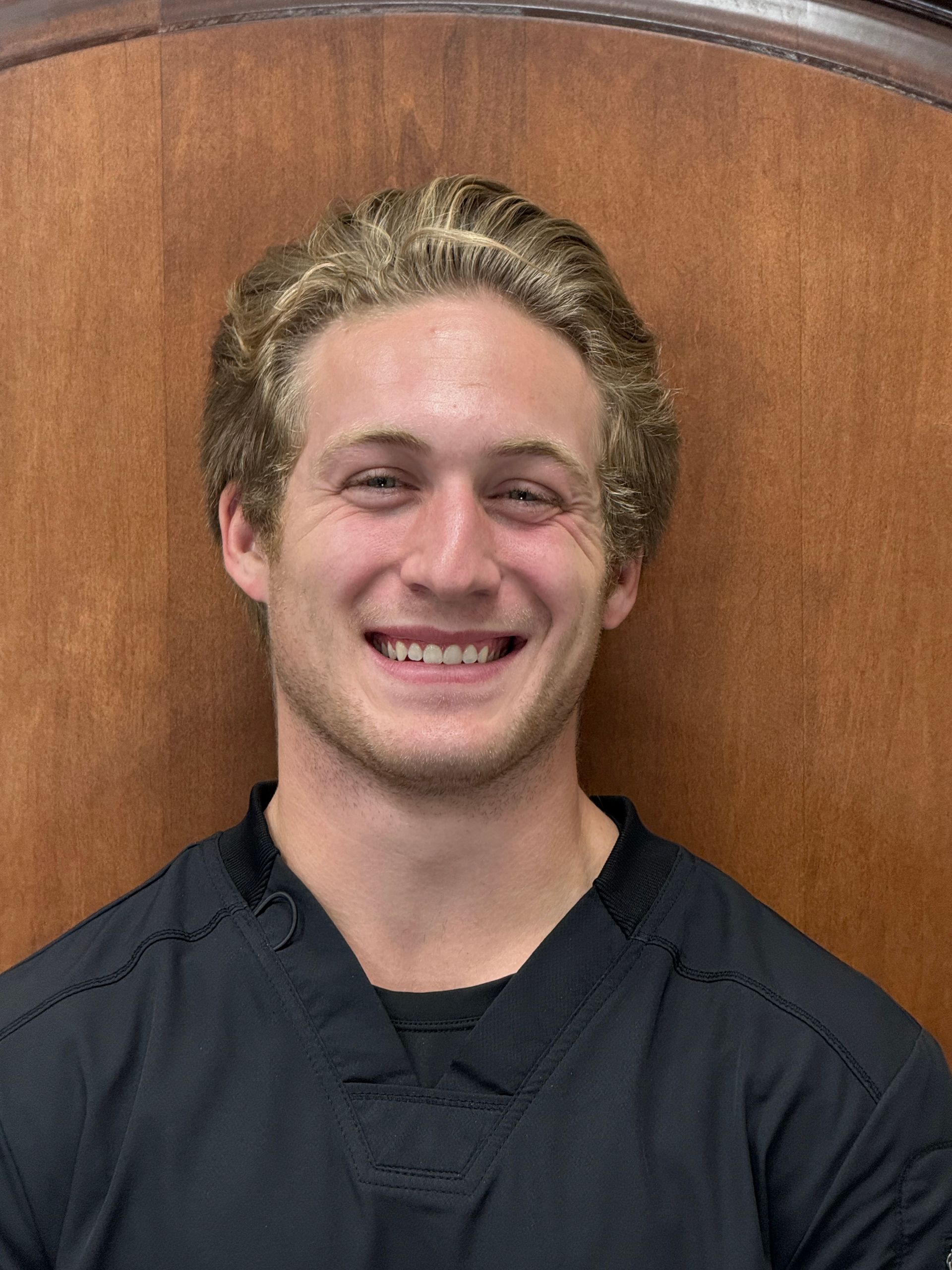 Smiling man with blonde hair and a black top against a wooden backdrop.