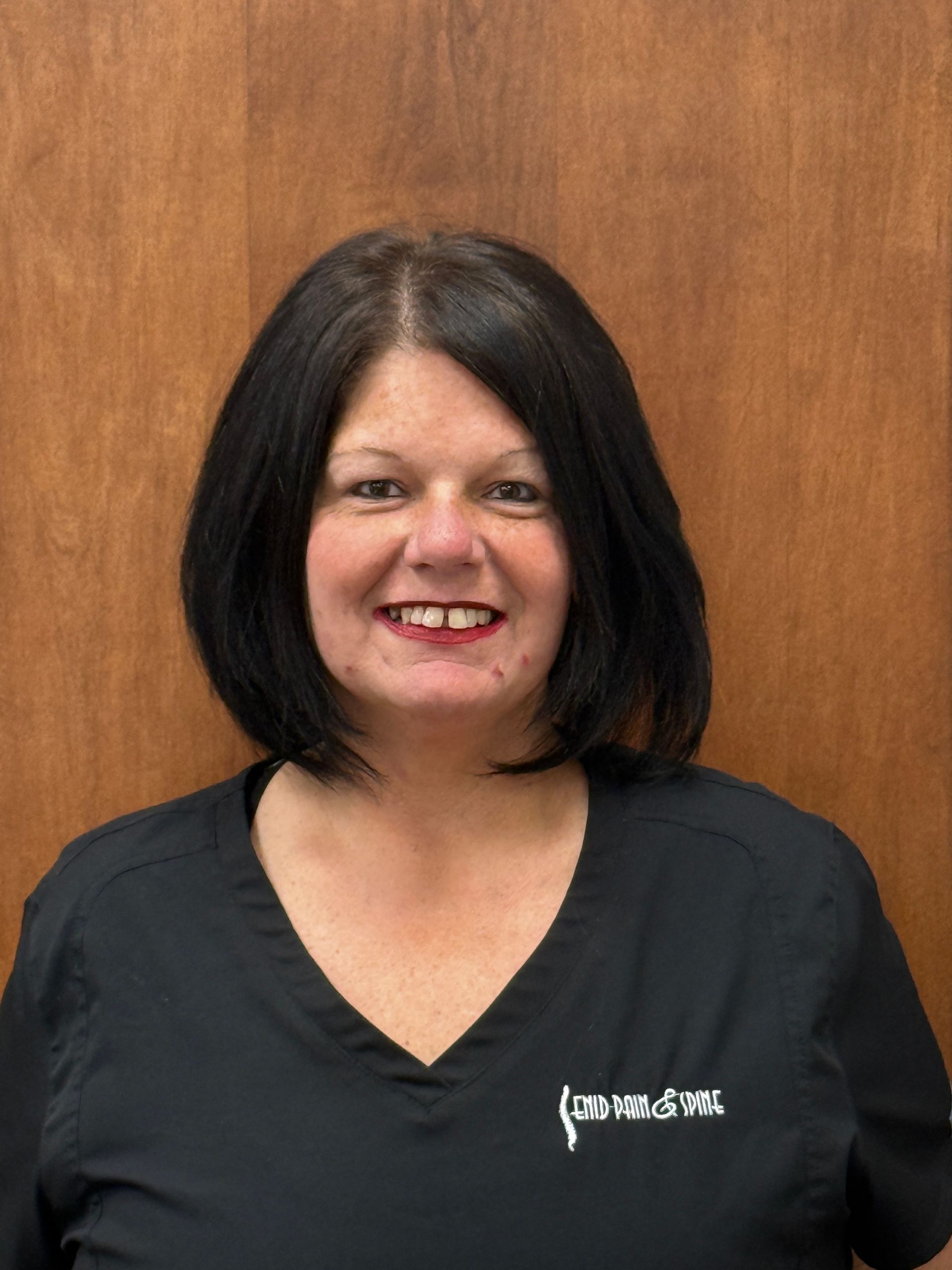 Woman with dark hair in a black v-neck shirt smiles, standing against a wood-paneled wall.
