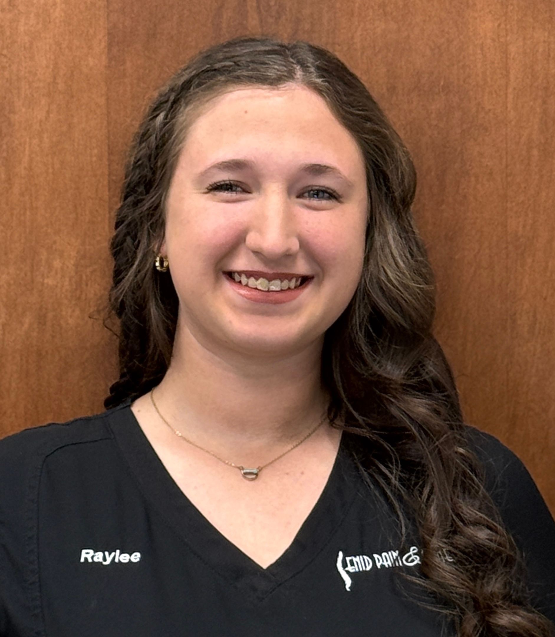 Smiling woman with long wavy hair, wearing a black top. Raylee is embroidered on the shirt. Wood background.